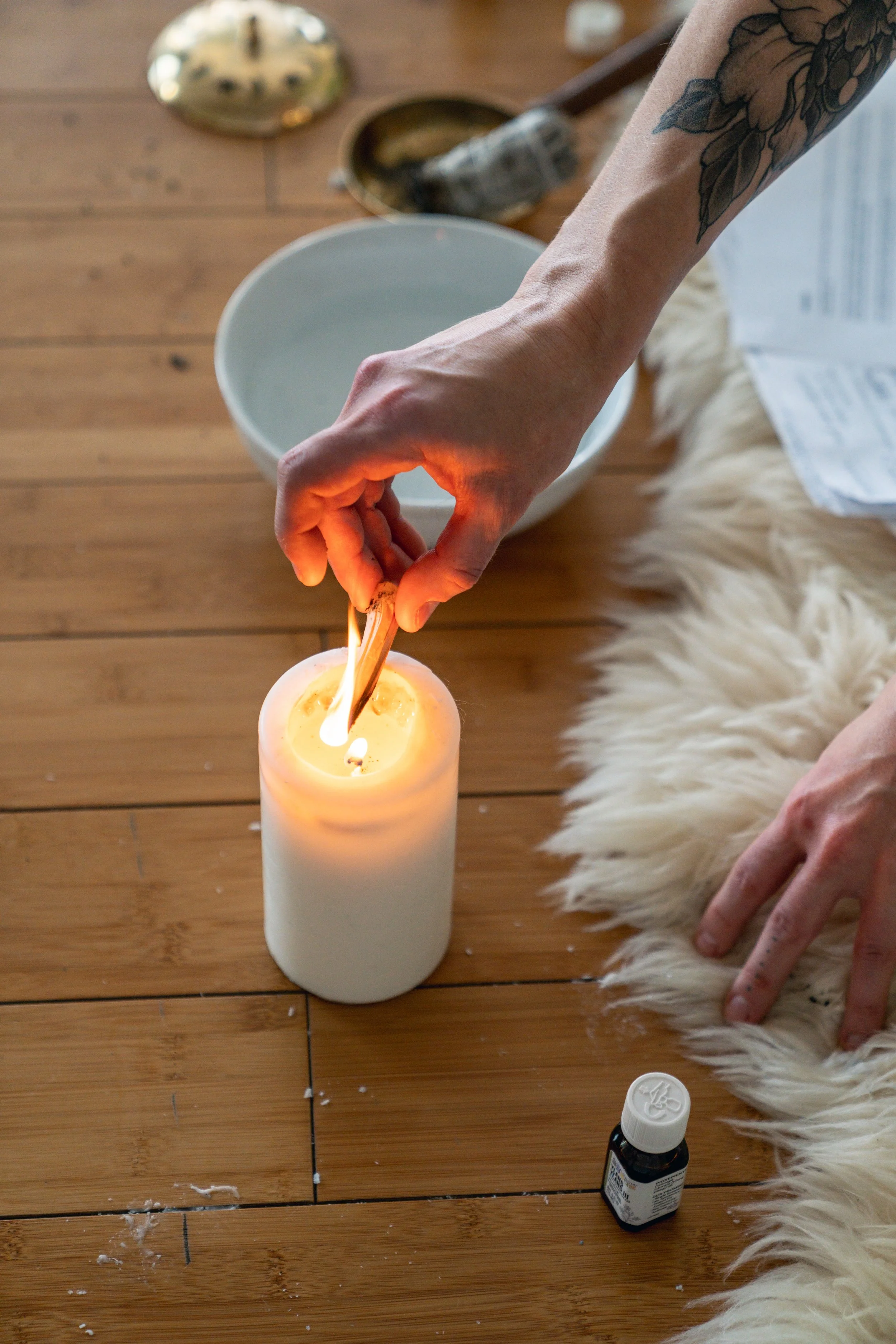 A person is lighting candles in a white pillar candle holder using a wooden matchstick. The scene takes place on a wooden floor with a fluffy white rug, a bowl, a small bottle, and some scattered items nearby.