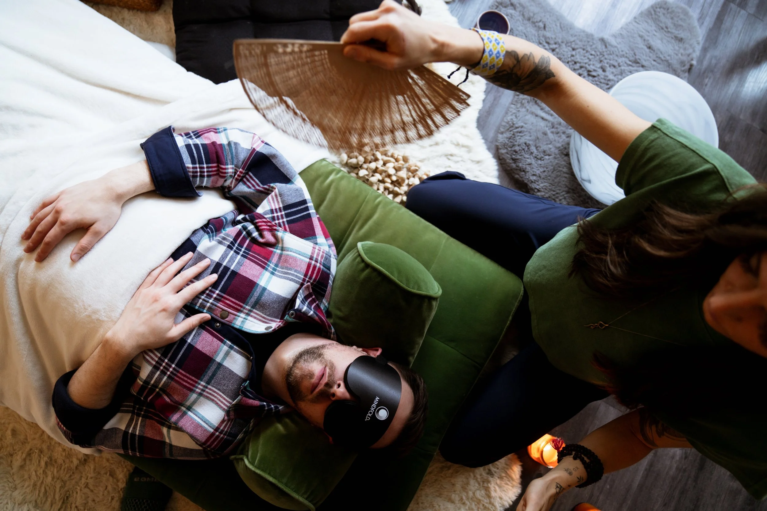 A man wearing an eye mask and plaid shirt lies on a green couch with his hand on his chest, while a woman in a green shirt sits nearby holding a fan and a candle on the floor. The scene is of a Breathwork Session, a relaxing or meditative activity.