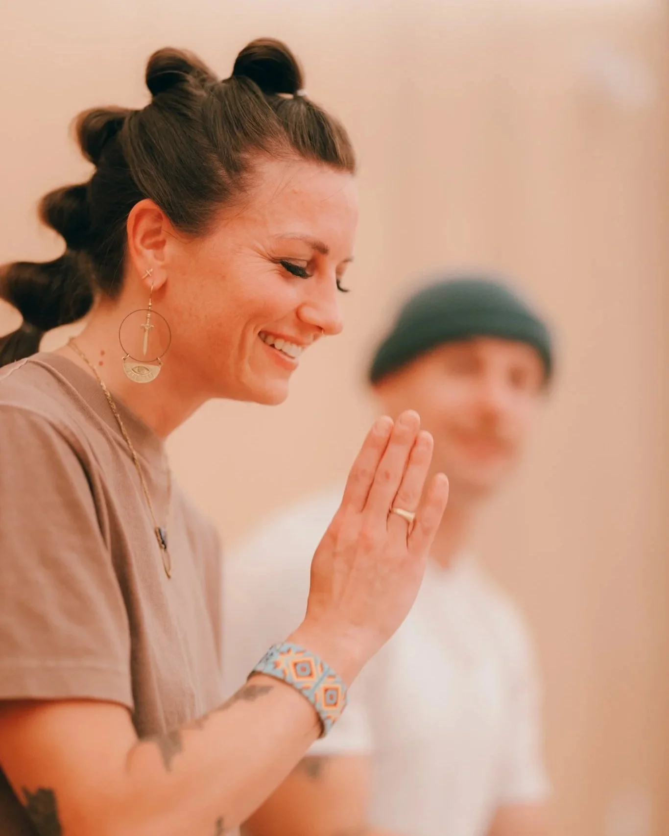 A woman with a joyful expression, eyes closed, smiling and holding her right hand up as if in prayer or gratitude. She has dark hair styled in rollers, is wearing long earrings, a watch, and a ring. A man wearing a beanie is blurred in the background.