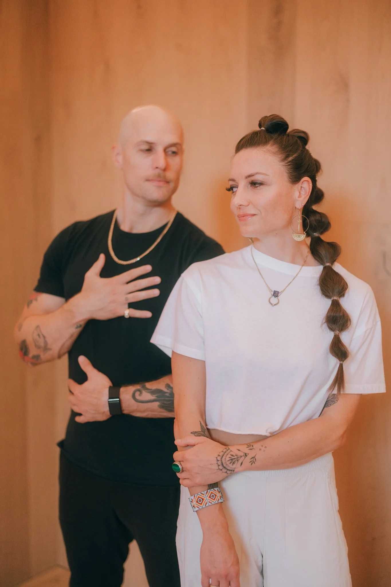 A man and a woman standing close together with a wooden background. The woman has a braid, tattoos on her arms, and is wearing a white T-shirt and jewelry. The man has tattoos, a shaved head, and is wearing a black T-shirt and a gold chain.