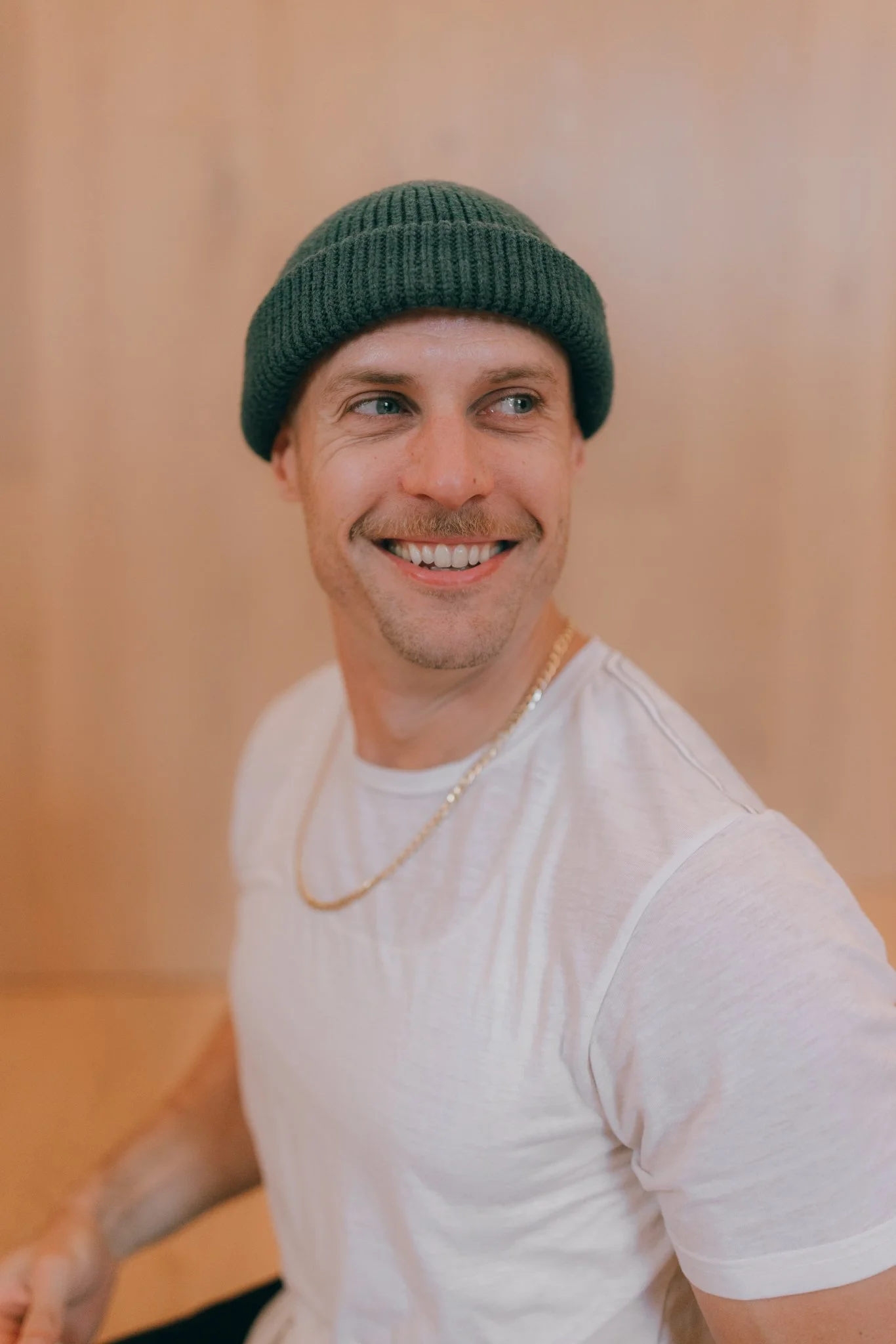 A young man, wearing a dark green knit beanie, a white t-shirt, a gold chain, and smiling while looking to the side.