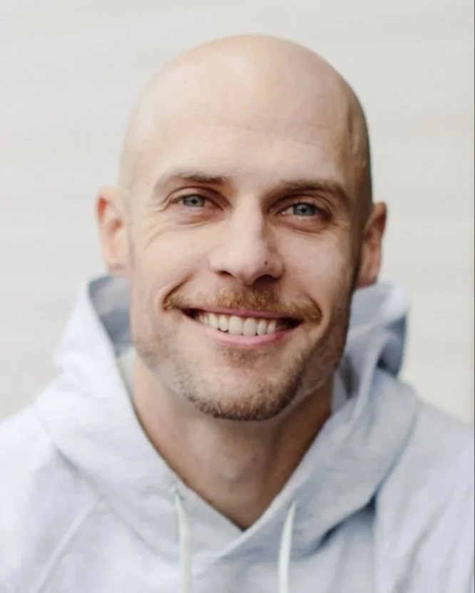 Close-up of smiling bald man wearing a light gray hoodie against a plain background.
