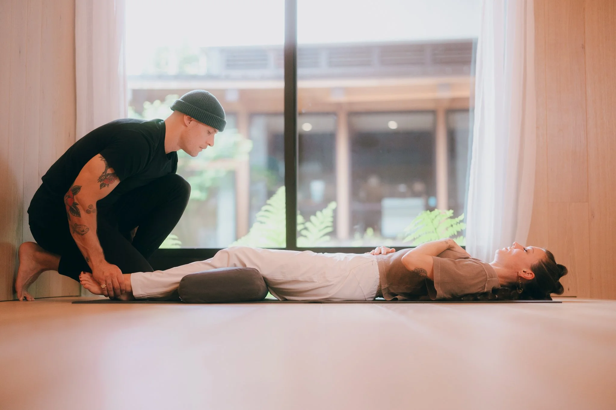 A person lying on their back with eyes closed, receiving a body posture adjustment from a man kneeling beside them, indoors near a large window with sheer curtains and a wooden wall.