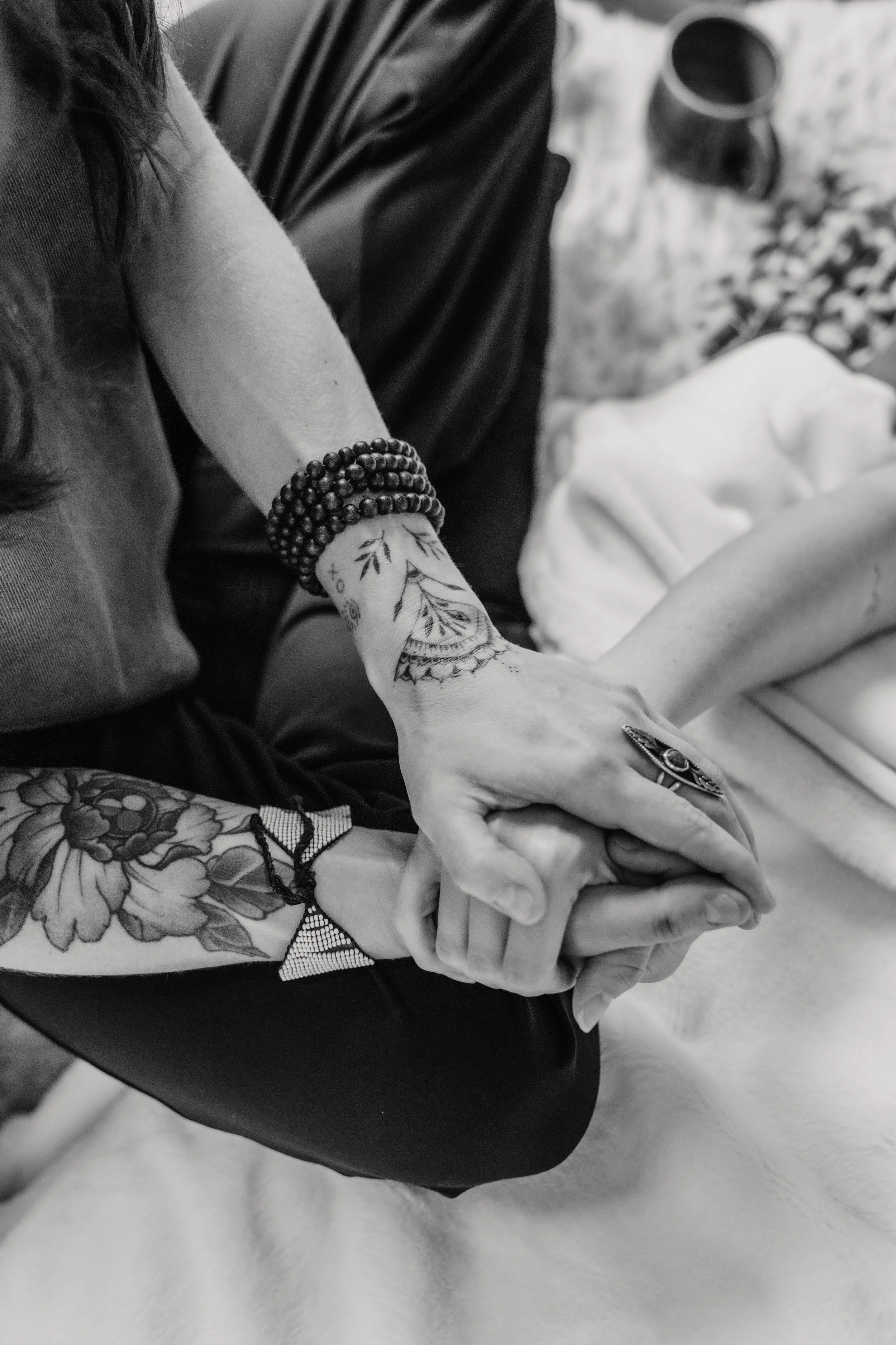 Black and white close-up of two people holding hands, showing tattoos and jewelry.