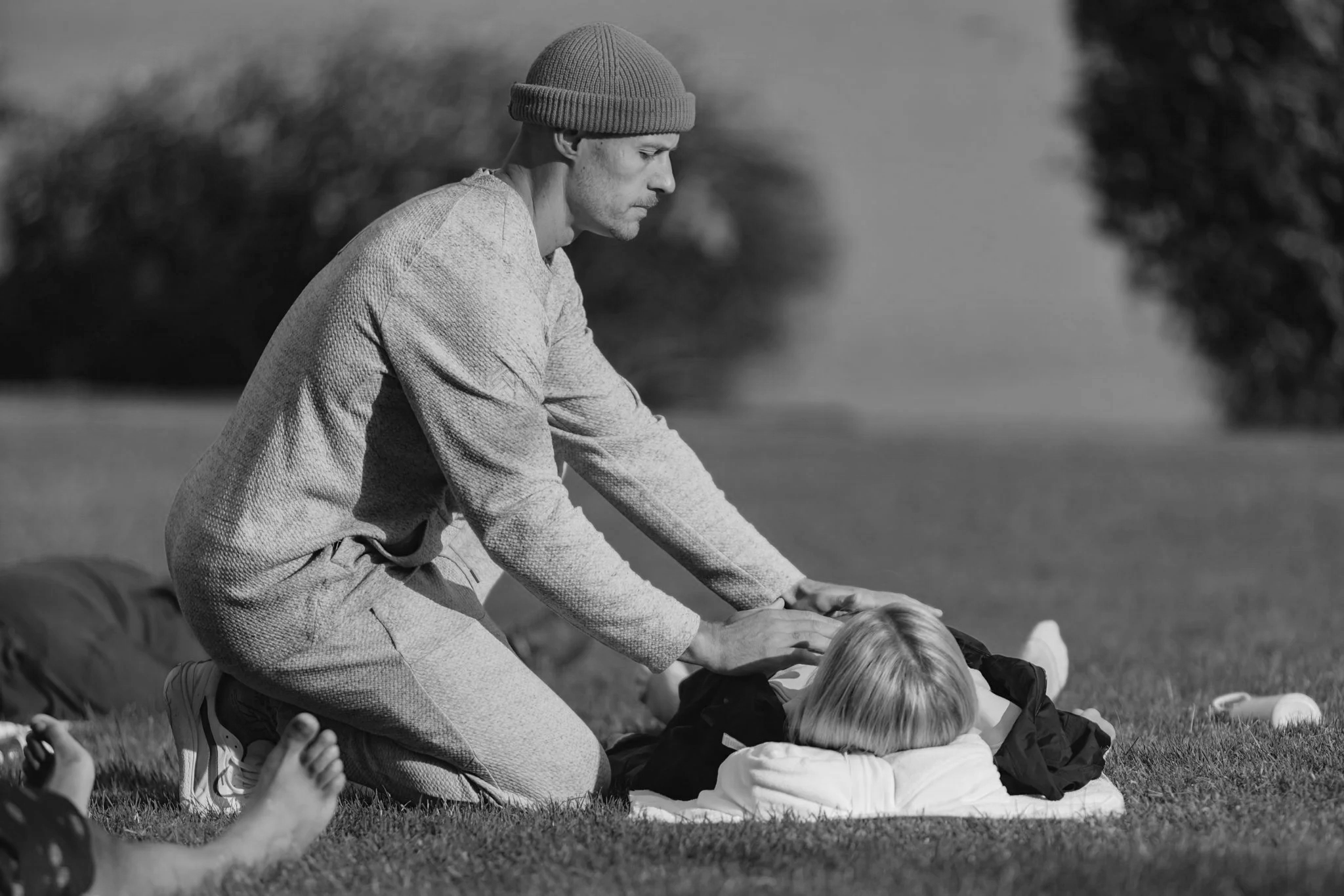 A man gives yoga body adjustments to a person lying on a blanket outdoors with trees and open space in the background.