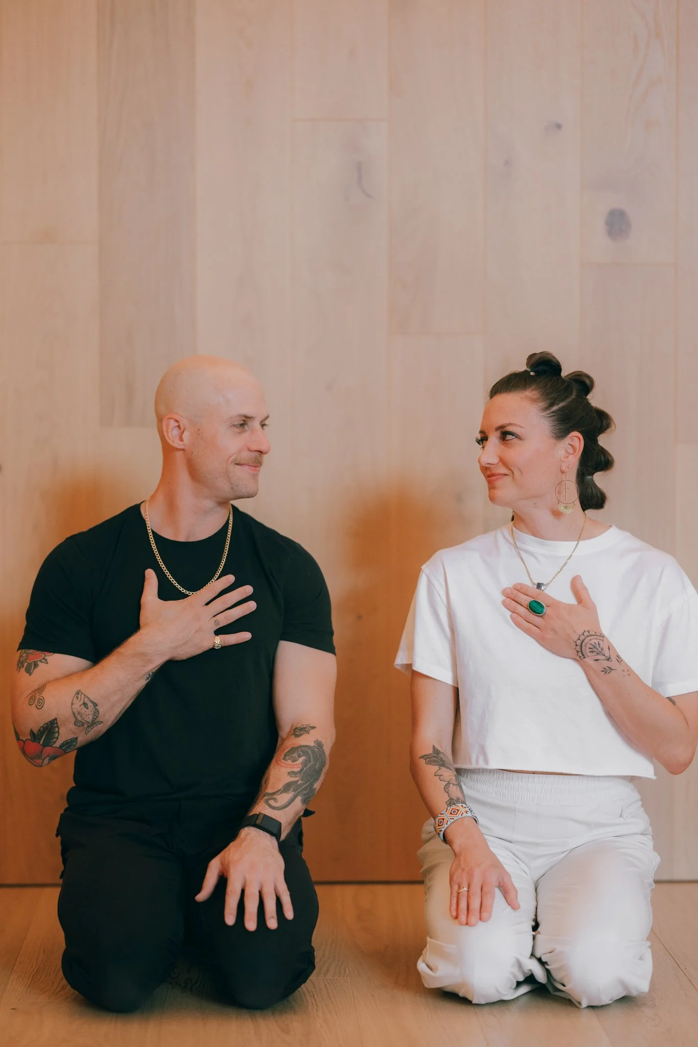 A man and woman sitting on the floor with wooden panel background, each with their right hand on their chest, smiling at each other.