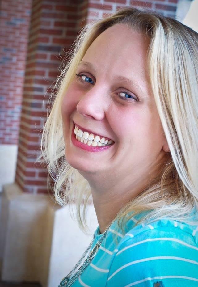 A smiling woman with blonde hair and blue eyes, wearing a blue striped shirt, sitting indoors with a brick wall in the background.