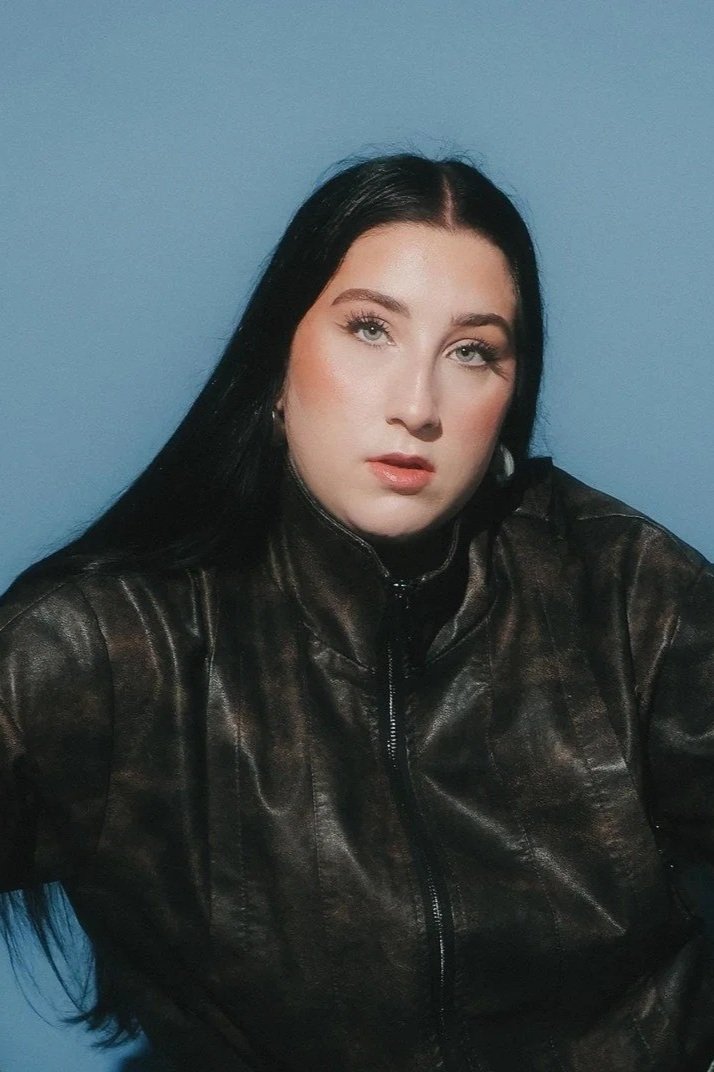 A woman with long black hair, wearing a black leather jacket, posing against a plain blue wall.