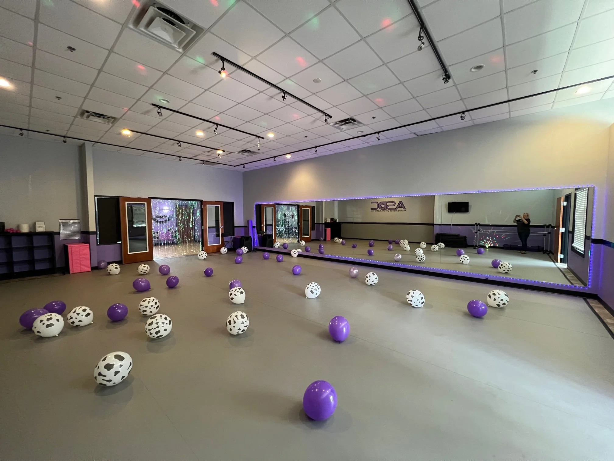 Empty dance studio with purple, black, and white balloons scattered on the floor, a large mirror along one wall, and a woman taking a photo in the reflection.