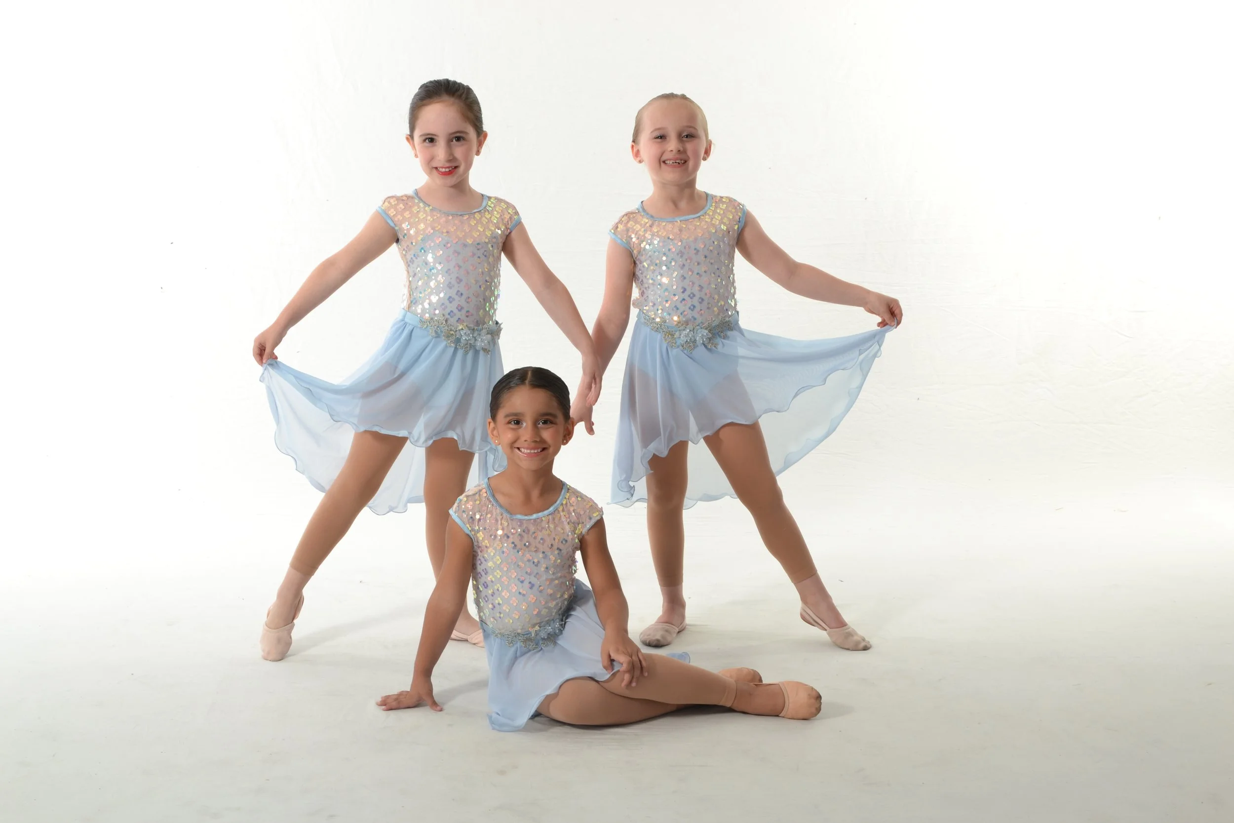 Three young girls in ballet costumes posing together against a plain white background, smiling at the camera.