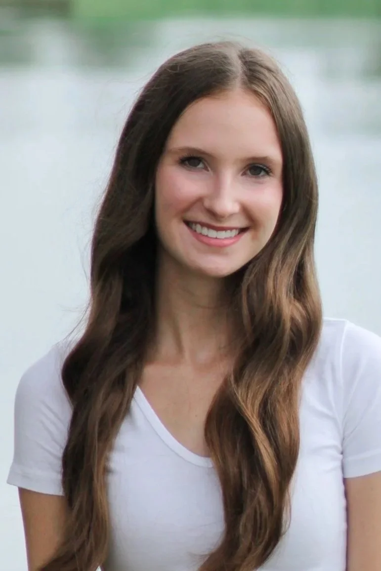 A young woman with long, wavy brown hair, smiling, standing outdoors near a body of water with greenery in the background, wearing a white t-shirt.