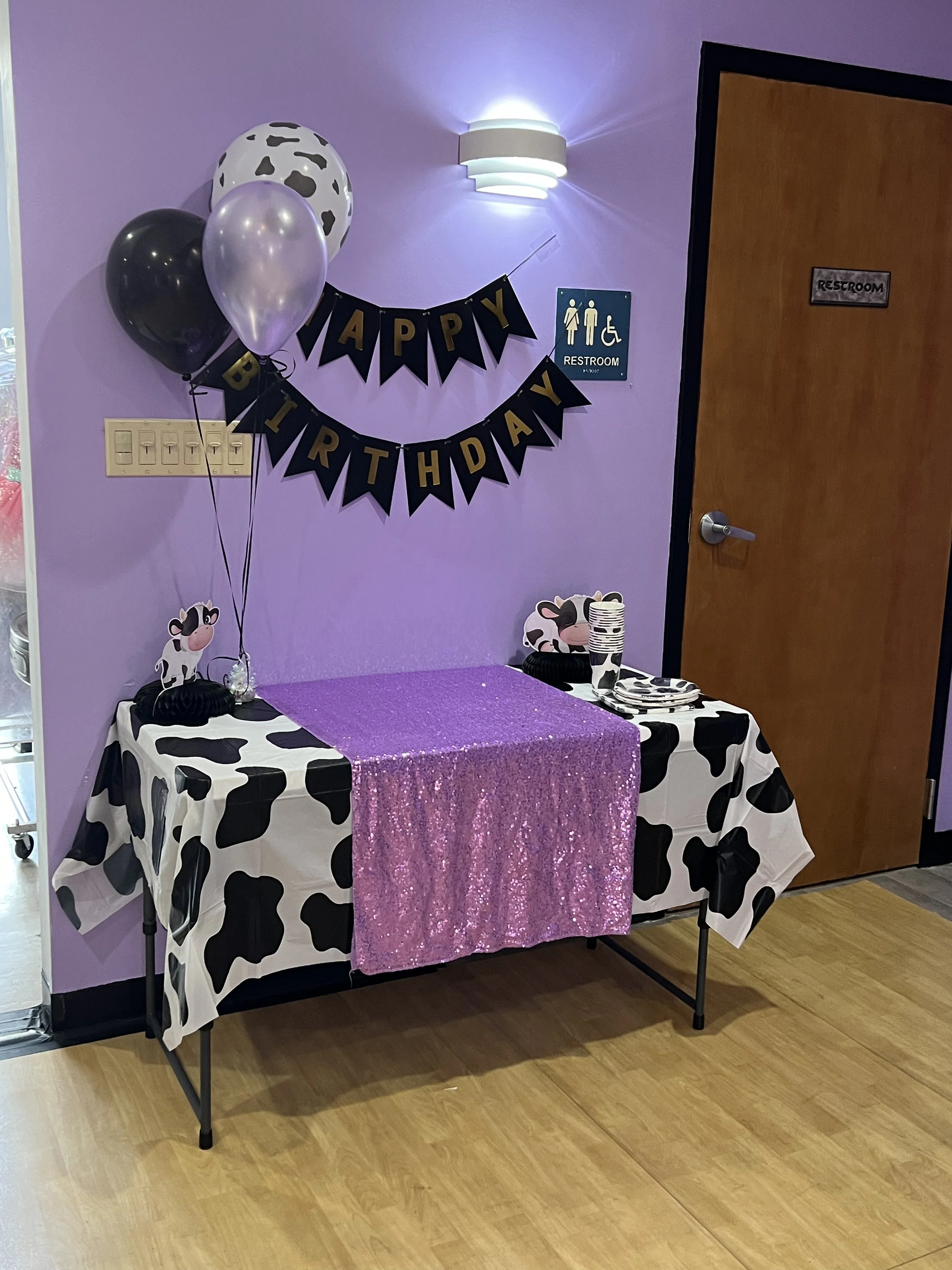 Cow-themed birthday party table with black and white cow print tablecloth, a pink shimmer table cover, cow-shaped decorations, balloons in cow print, light purple wall, and a 'Happy Birthday' banner.