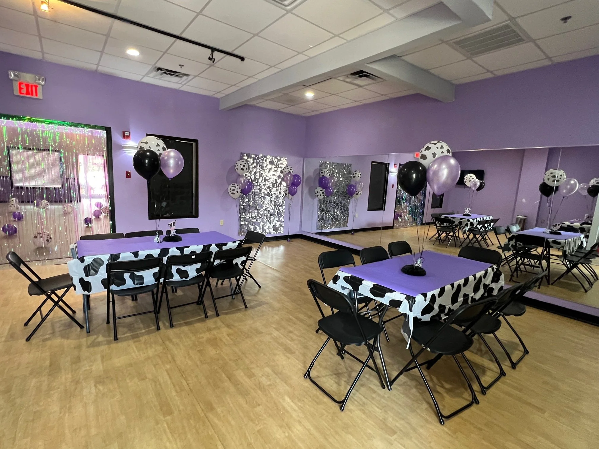 Decorated party room with purple walls, tables covered with cow-print tablecloths, black chairs, and black, purple, and white balloons. A mirrored wall and a reflective backdrop with hanging decorations are visible.