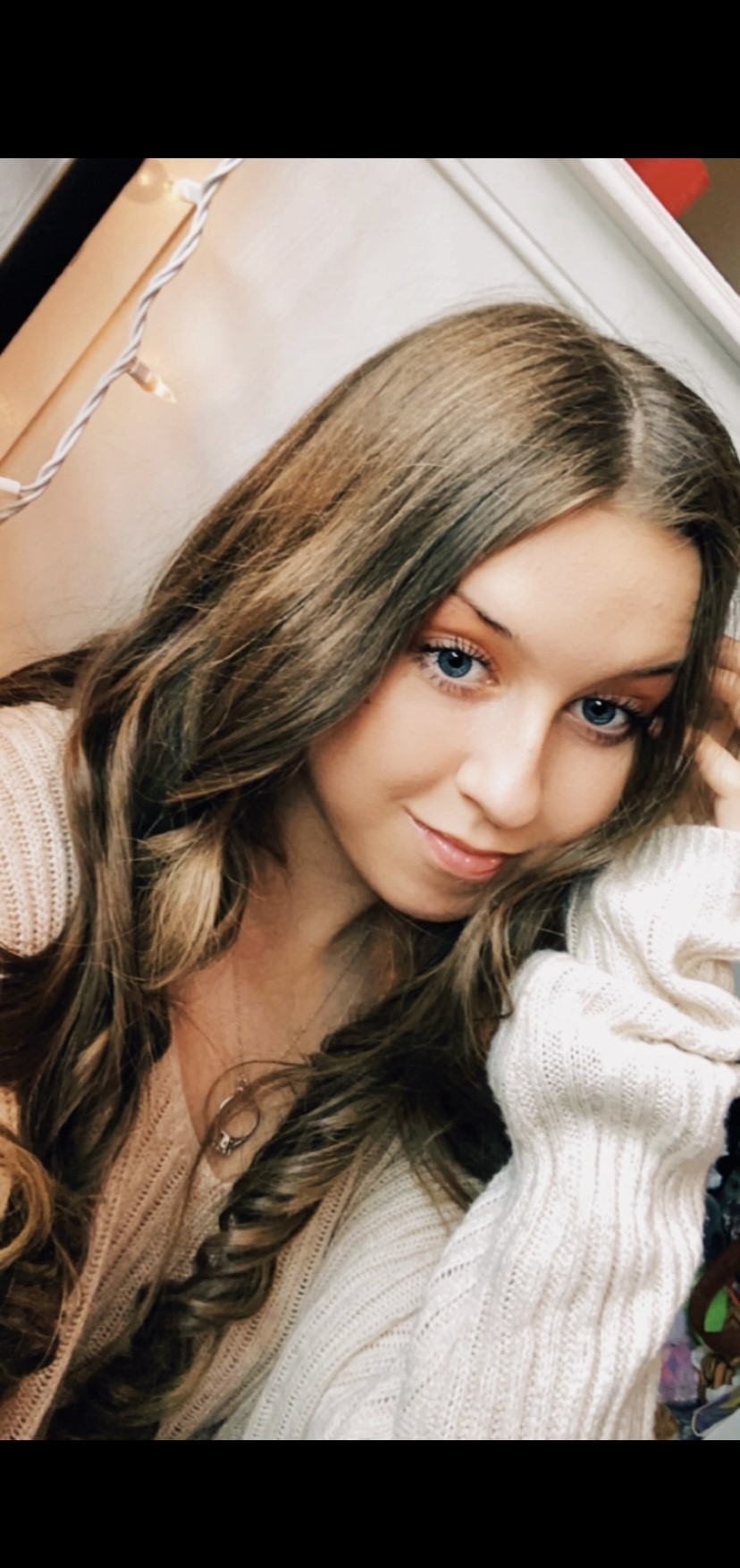 Close-up of a young woman with long, wavy brown hair and blue eyes, wearing a cream-colored knit sweater, smiling softly at the camera.