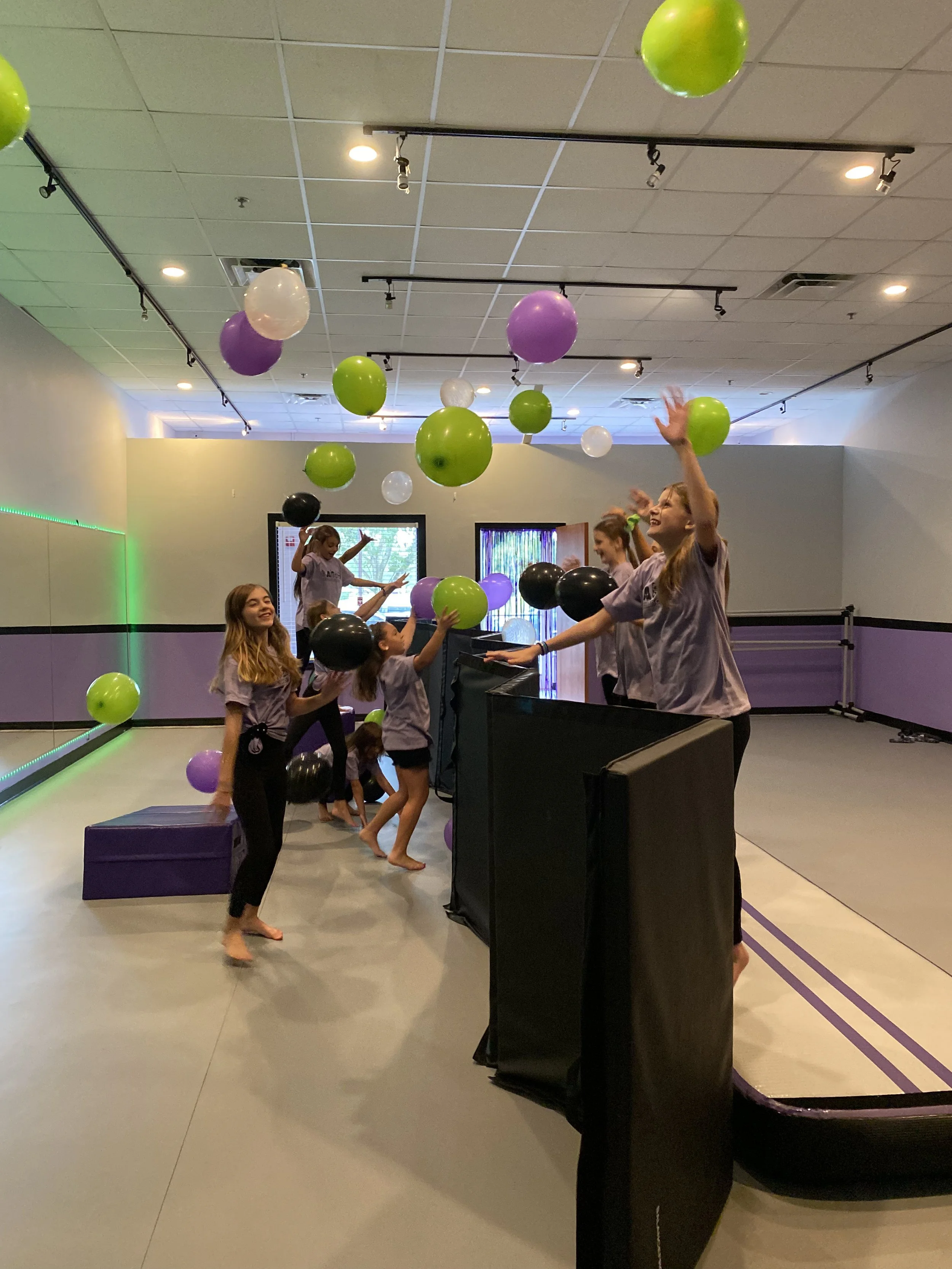 A group of young girls in matching purple shirts playing with black, green, purple, and white balloons in an indoor gym with mirrored walls and a padded area.
