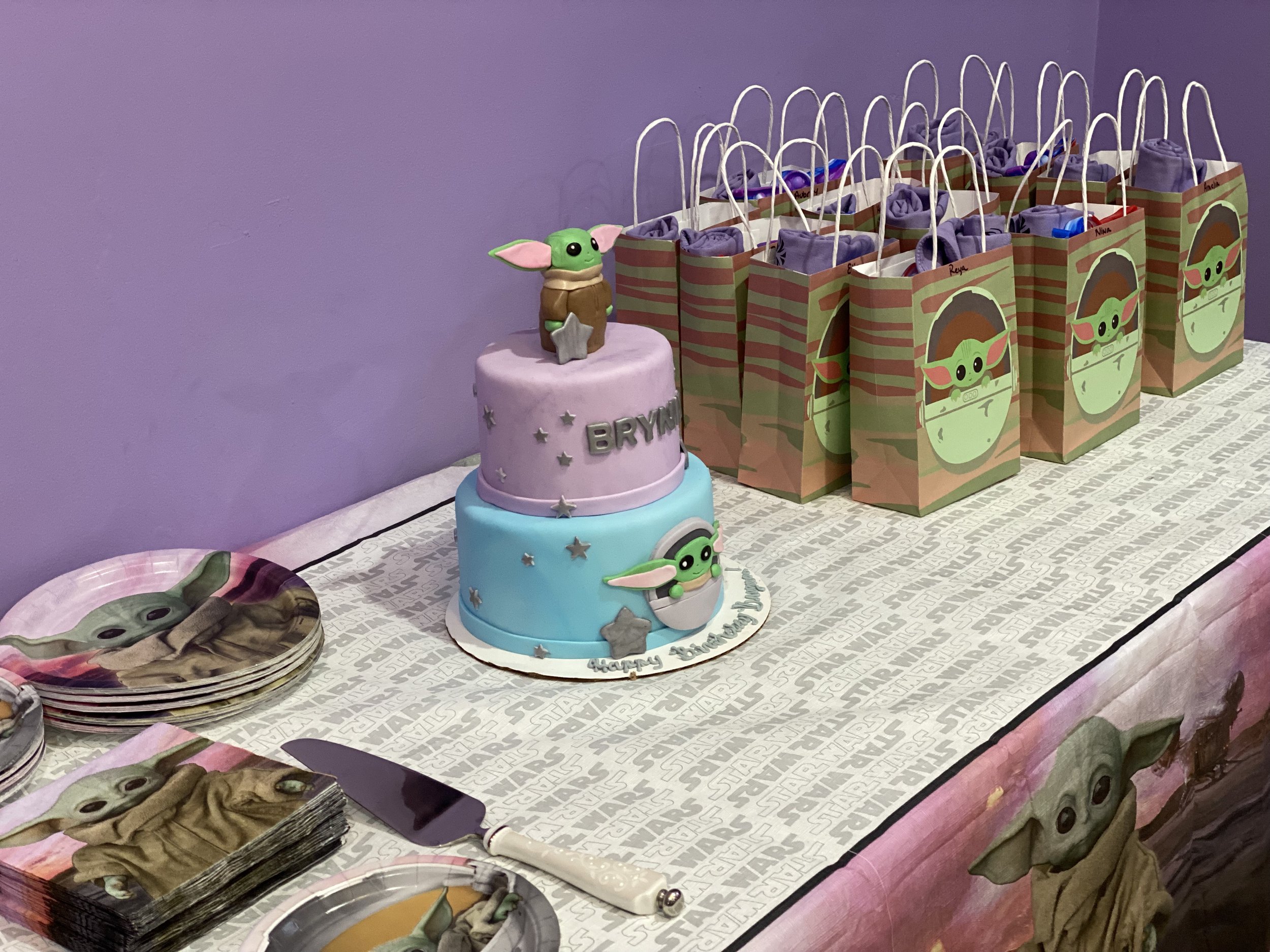 Star Wars themed birthday party table with a two-tier cake featuring Baby Yoda (Grogu) decorations, plates with Baby Yoda images, and party favor bags also decorated with Baby Yoda on a purple wall background.