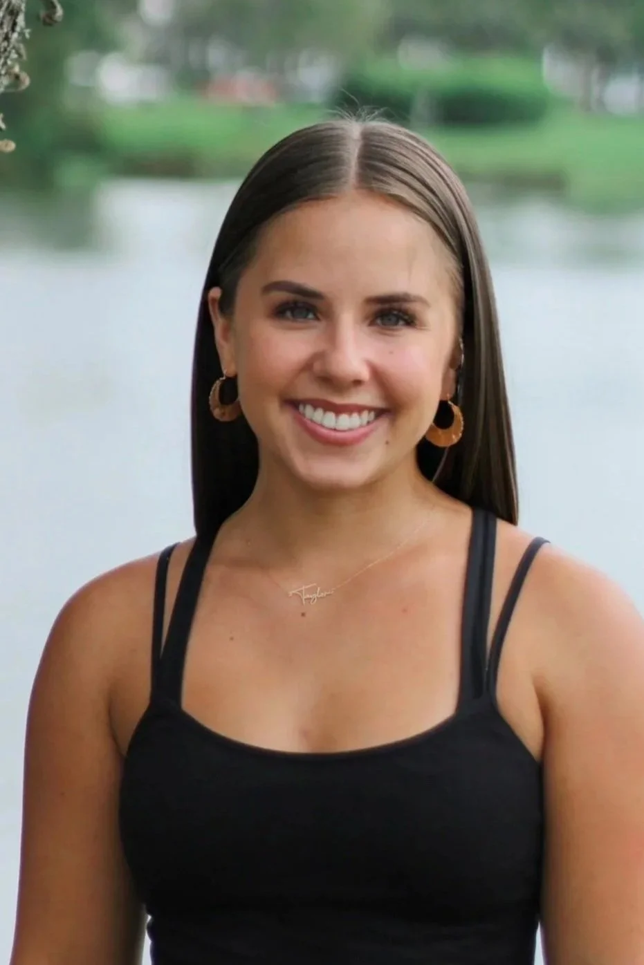 A young woman with long brown hair, wearing a black spaghetti strap top, gold hoop earrings, and a delicate necklace, smiling outdoors near a body of water with greenery in the background.