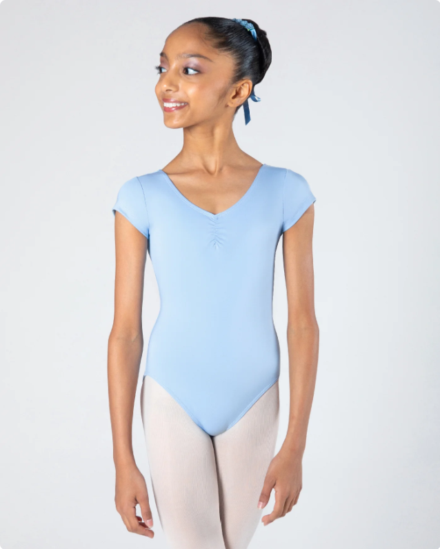Young girl in light blue fitness leotard and pink leggings smiling and looking to the side, with a blue hair ribbon, standing against a plain light background.