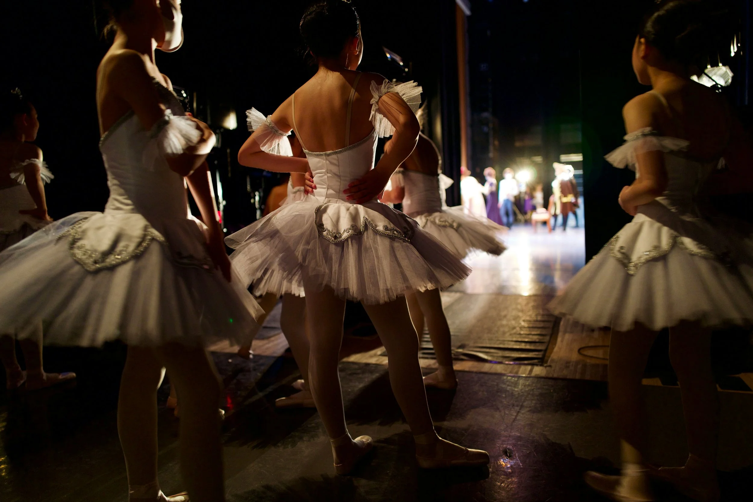 Ballet dancers in tutu costumes backstage, preparing to perform, with stage visible in the background.