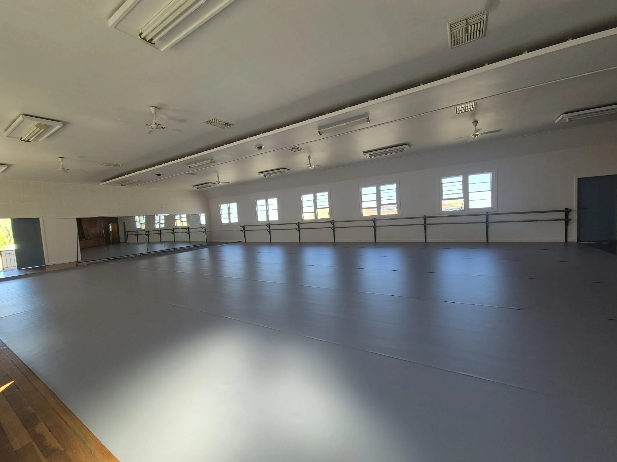 Interior of a dance studio with a large open floor, mirrors on the wall, ballet barres along the windows, and ceiling lights.