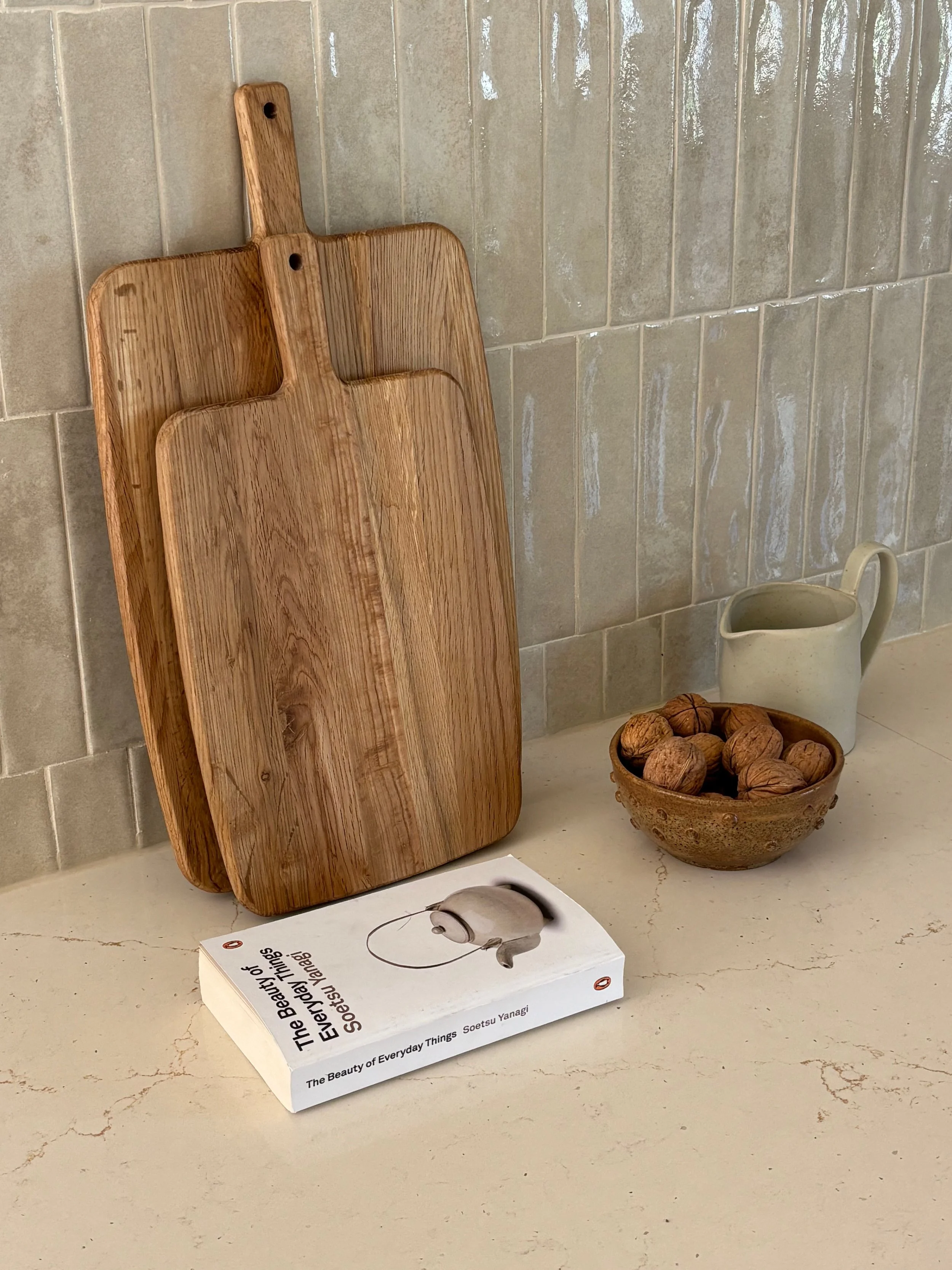 A kitchen countertop with two hanging wooden cutting boards, a white mug, a bowl of walnuts, and a book titled 'The Beauty of Everyday Things' by Soetsu Yanagi.