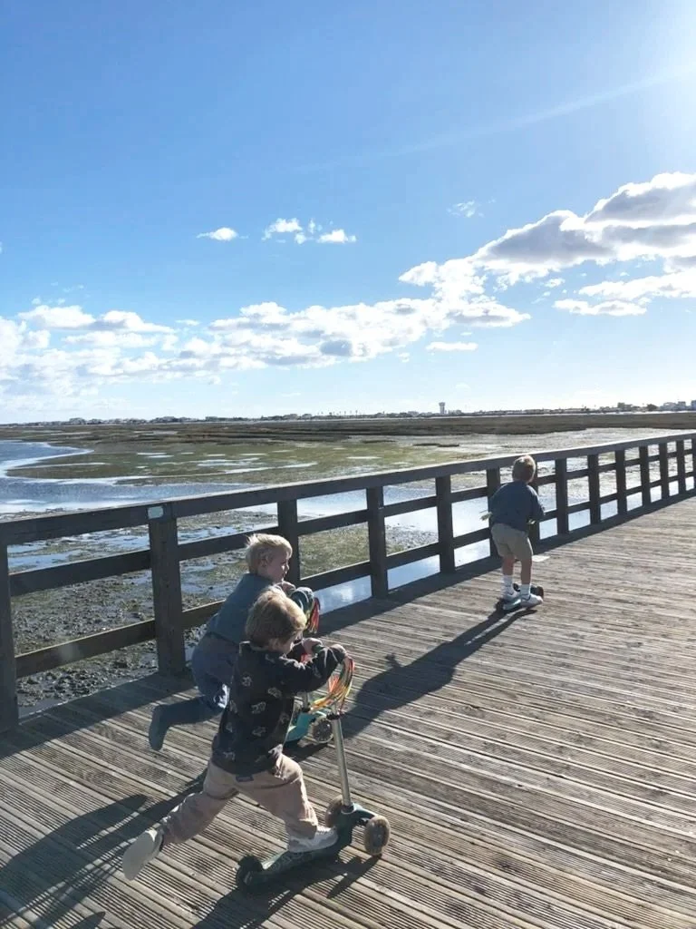 Three children playing on a wooden pier overlooking a marshy area and water with a cloudy sky overhead.