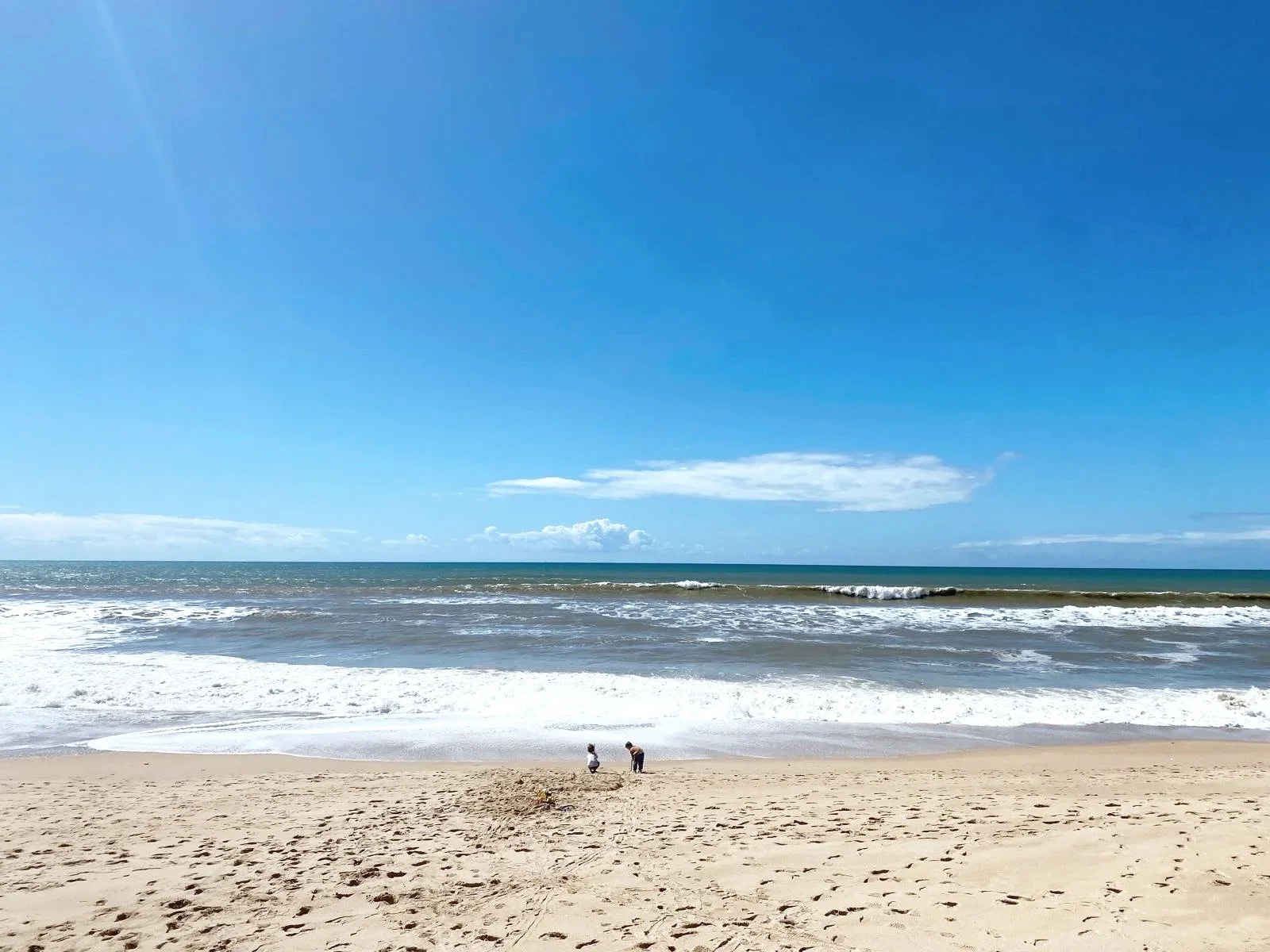A sandy beach with a few people near the shoreline, blue ocean waves, and a clear sky with some clouds.