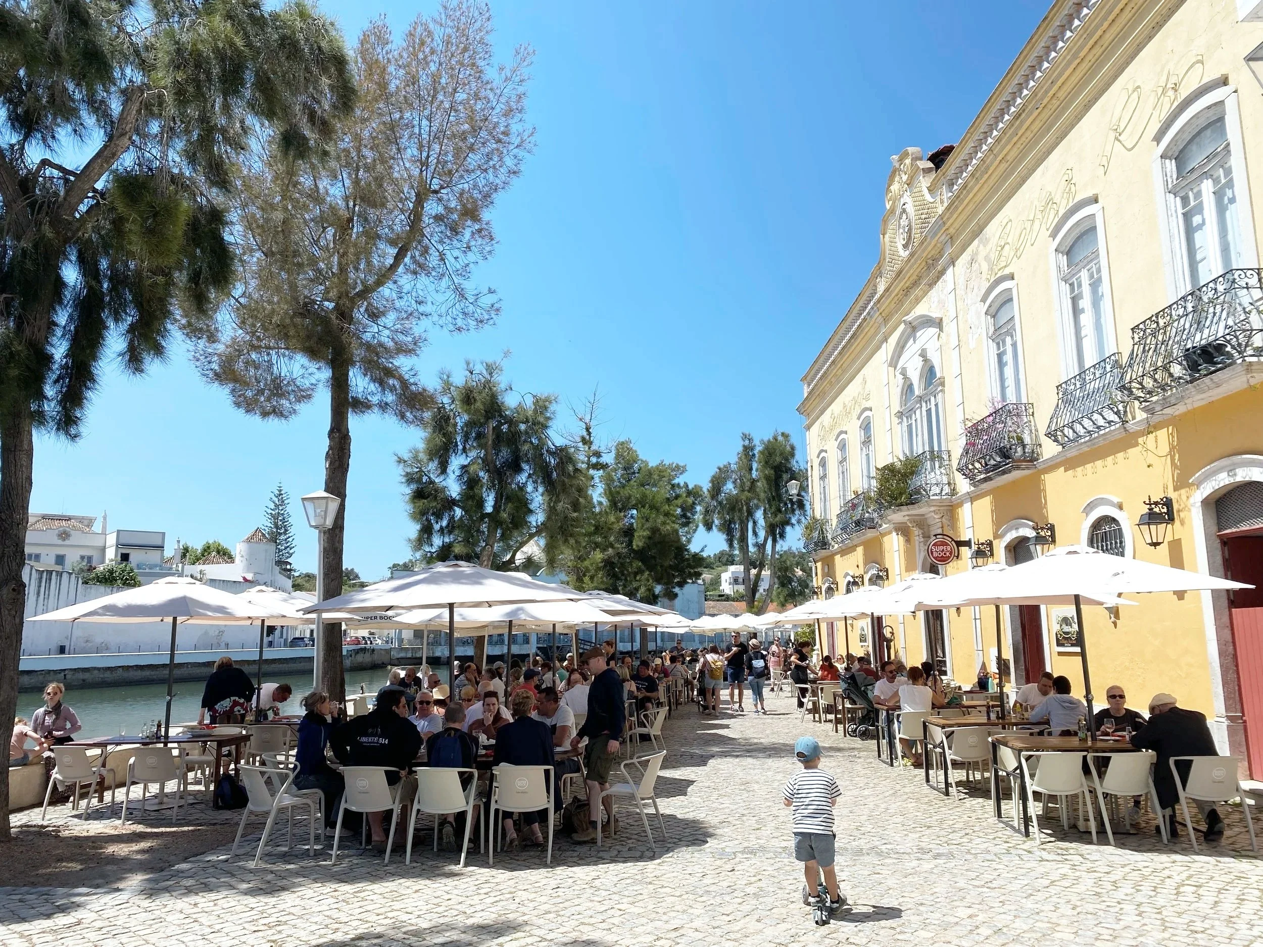 Outdoor cafe with people dining under umbrellas near a canal, yellow historic building, trees, and a clear blue sky.