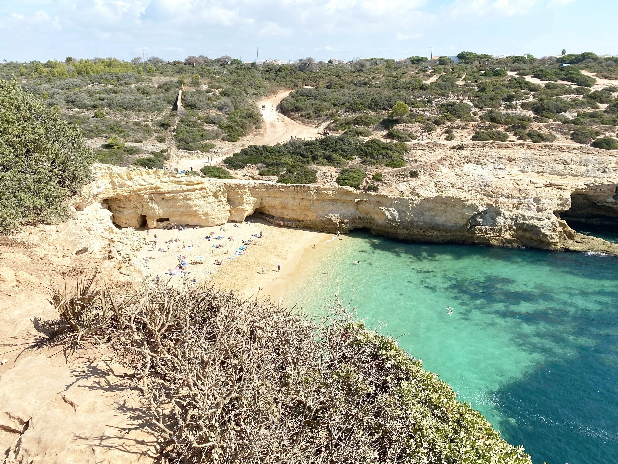 A beach with golden sand, turquoise water, and rocky cliffs, with some people sunbathing and swimming, surrounded by green bushes and hills under a partly cloudy sky.