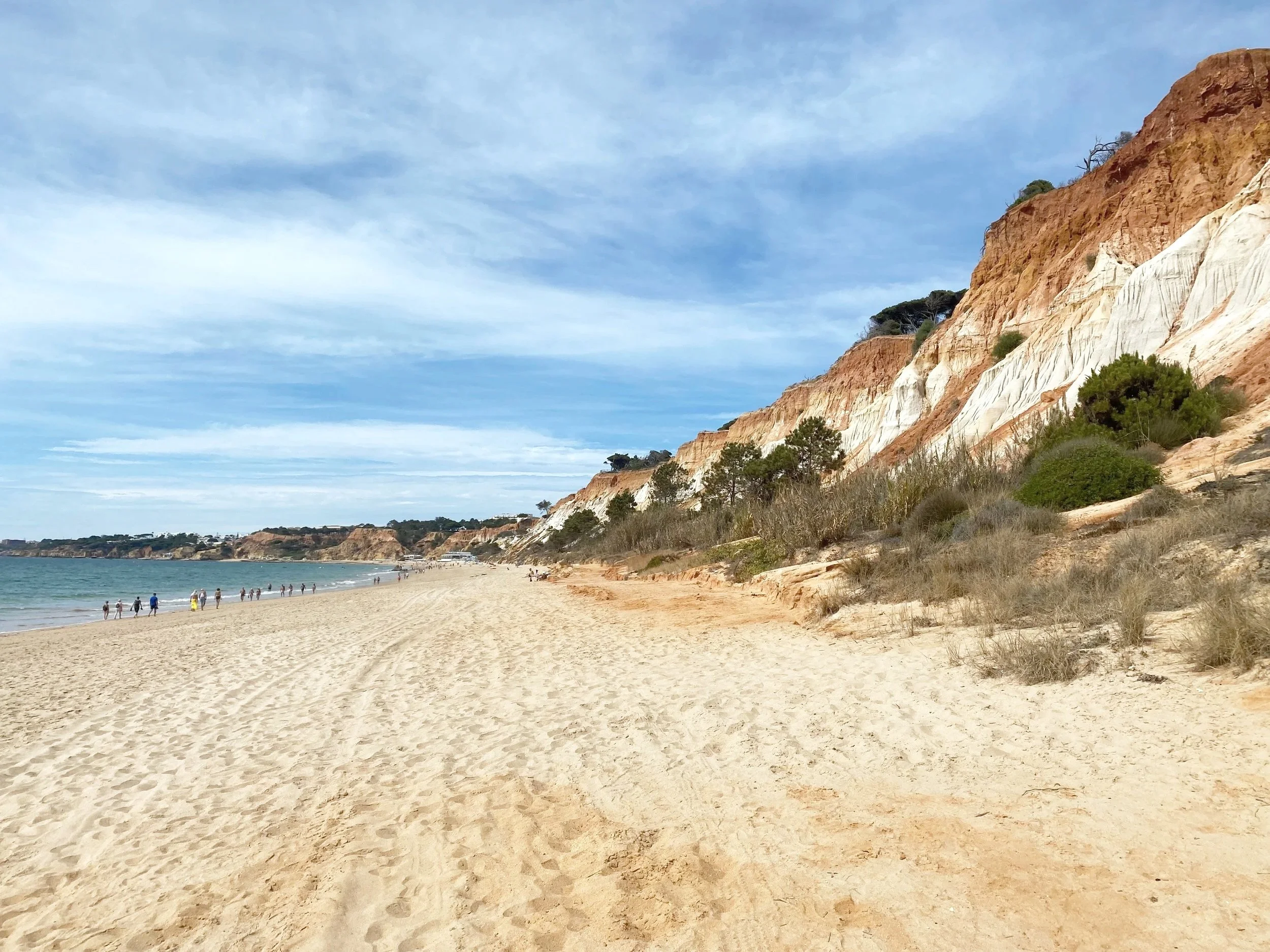 A sandy beach with a shoreline and cliffs in the background. People are walking along the beach under a partly cloudy sky.