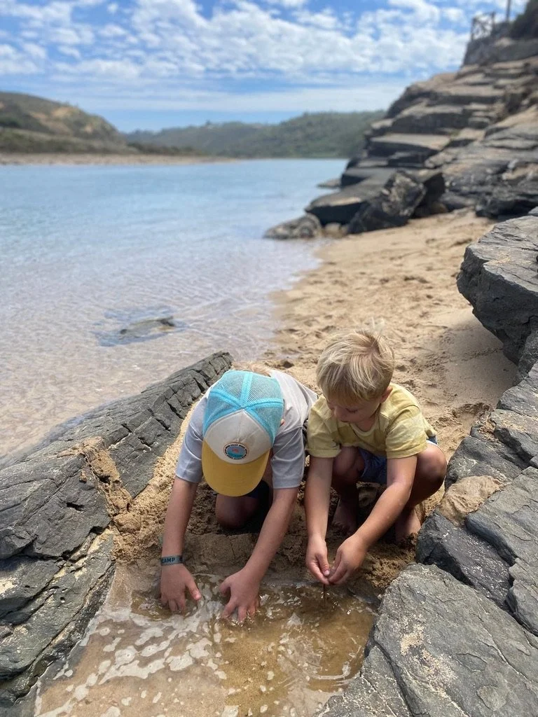 Two young boys playing by a small stream on a sandy beach with rocks, overlooking a body of water and distant hills under a partly cloudy sky.