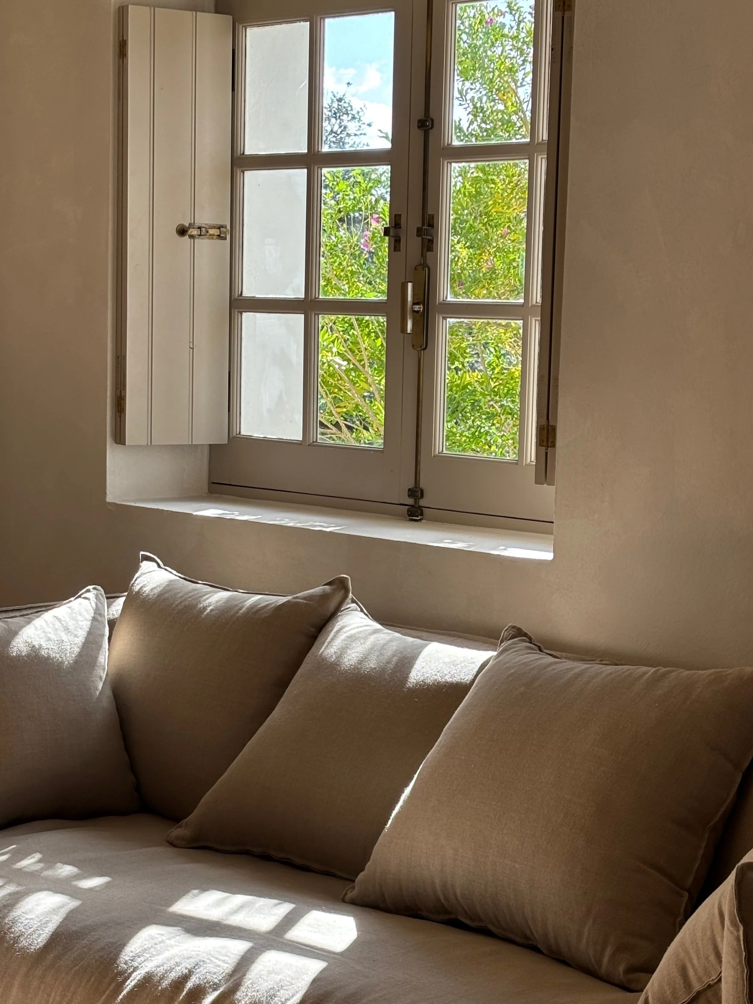 Sunlight streaming through a window with wooden shutters, illuminating a beige sofa with matching cushions.