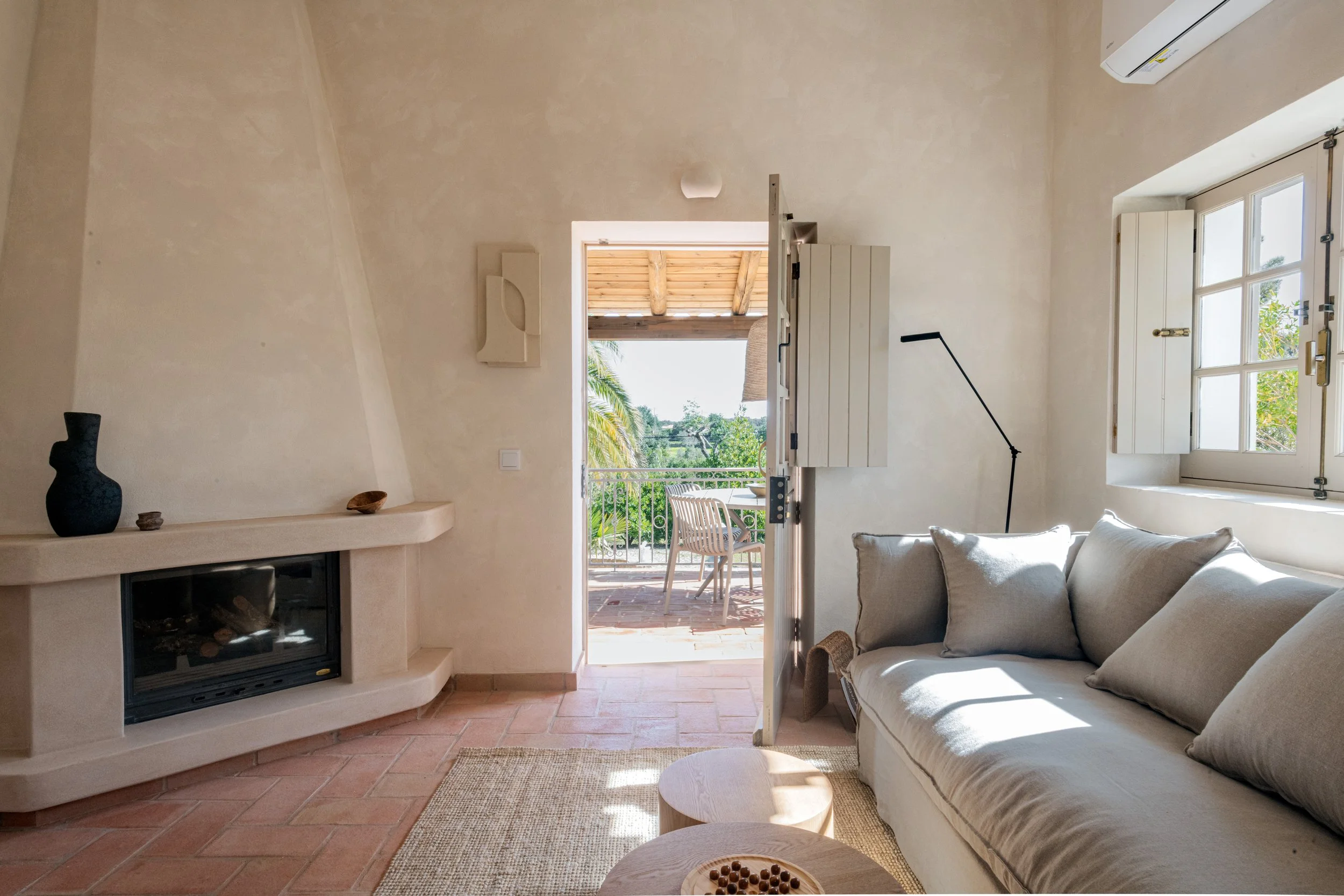 Living room with a beige sofa, a black floor lamp, a fireplace with decorative vases on the mantel, an open door leading to a balcony with outdoor furniture, a window with open shutters, and sunlight streaming into the room.