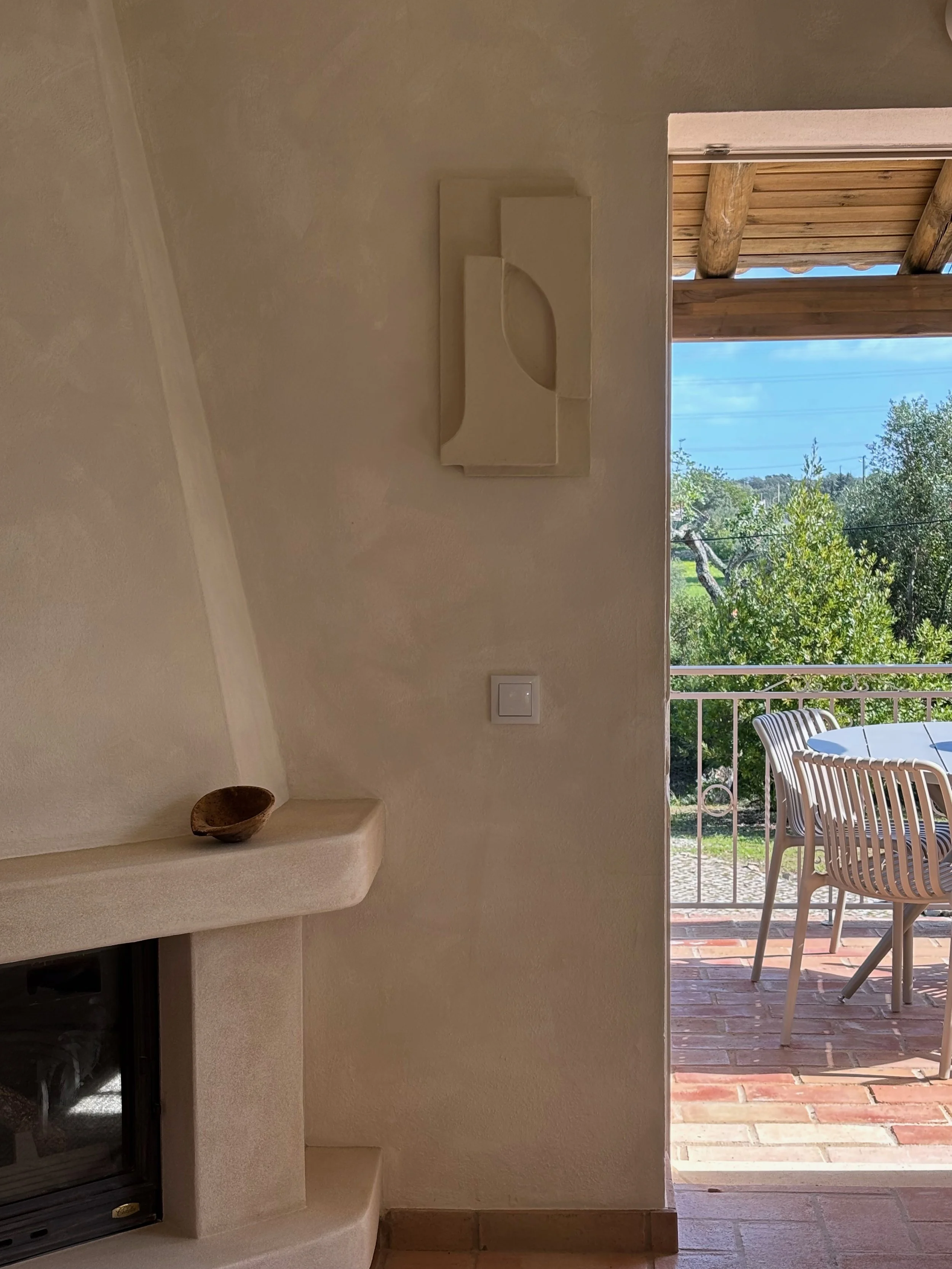 Interior view showing a fireplace with a small wooden bowl on the mantel, and an open door leading to a patio with outdoor chairs and a table, with trees and sky visible outside.