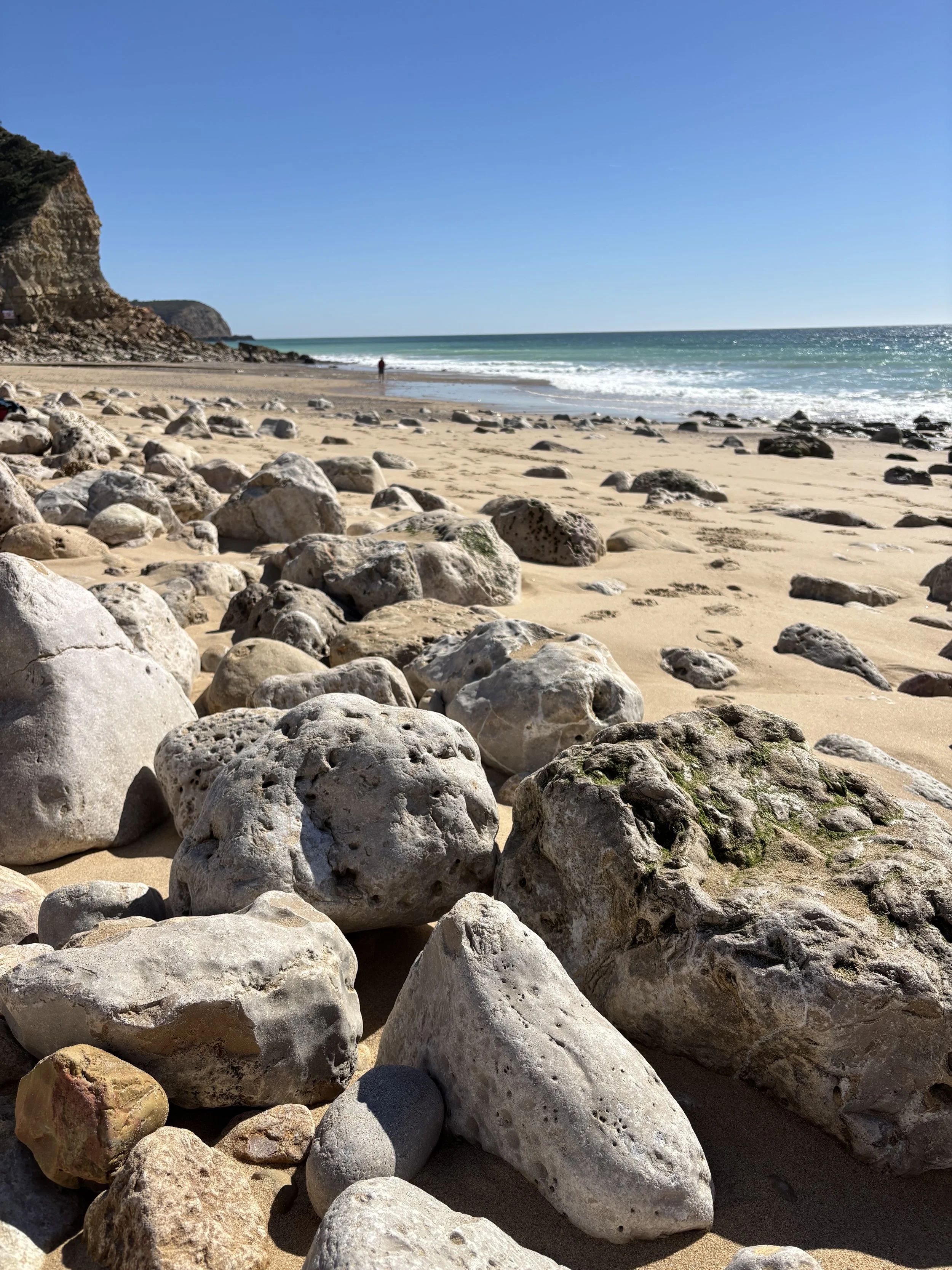 Rocky beach with sand, ocean waves, cliffs in the distance, and a clear blue sky.