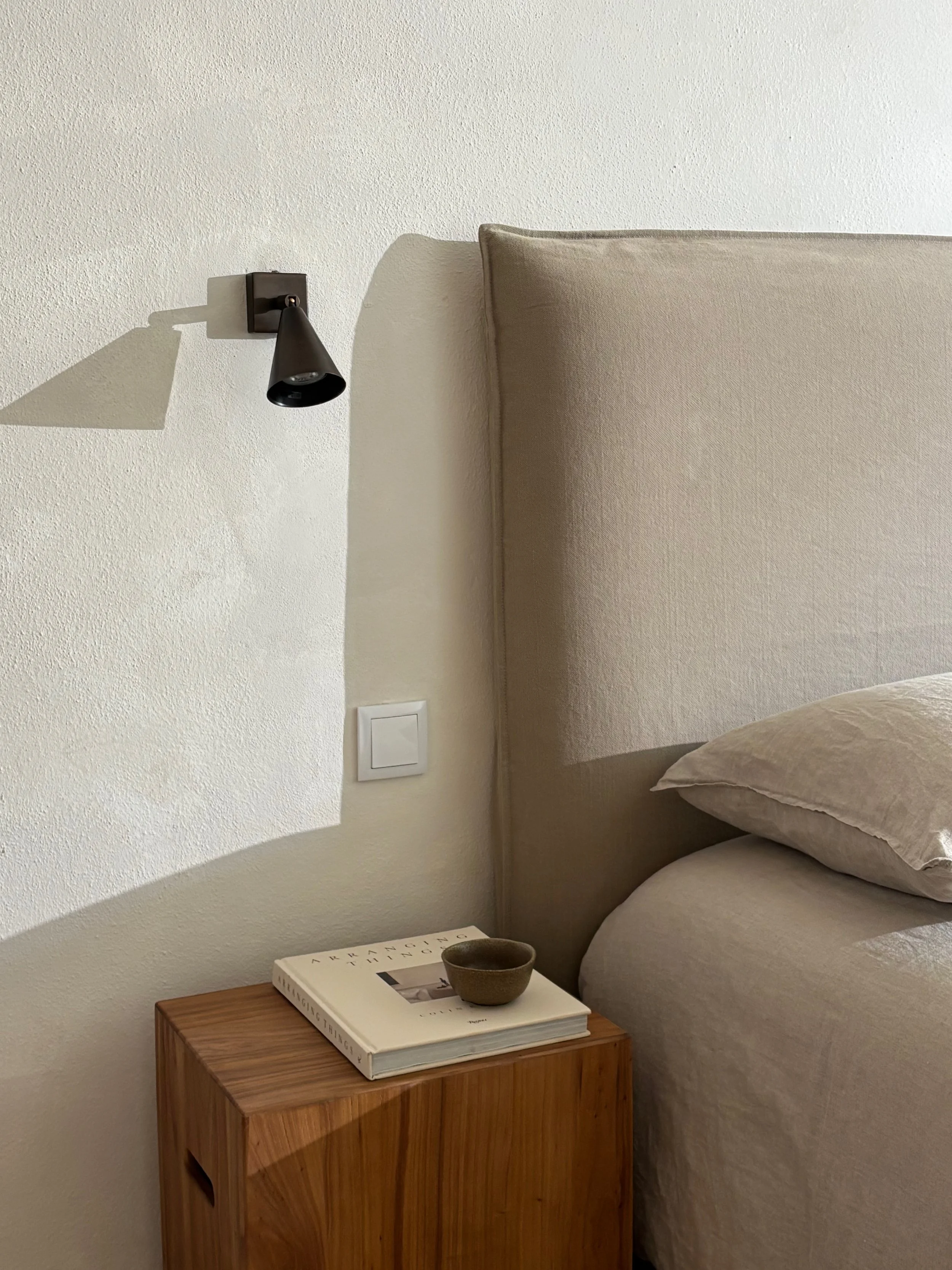 A bedroom corner with a beige upholstered headboard, a wooden nightstand with a book and a small ceramic bowl, a white textured wall with a black adjustable wall lamp casting a shadow.