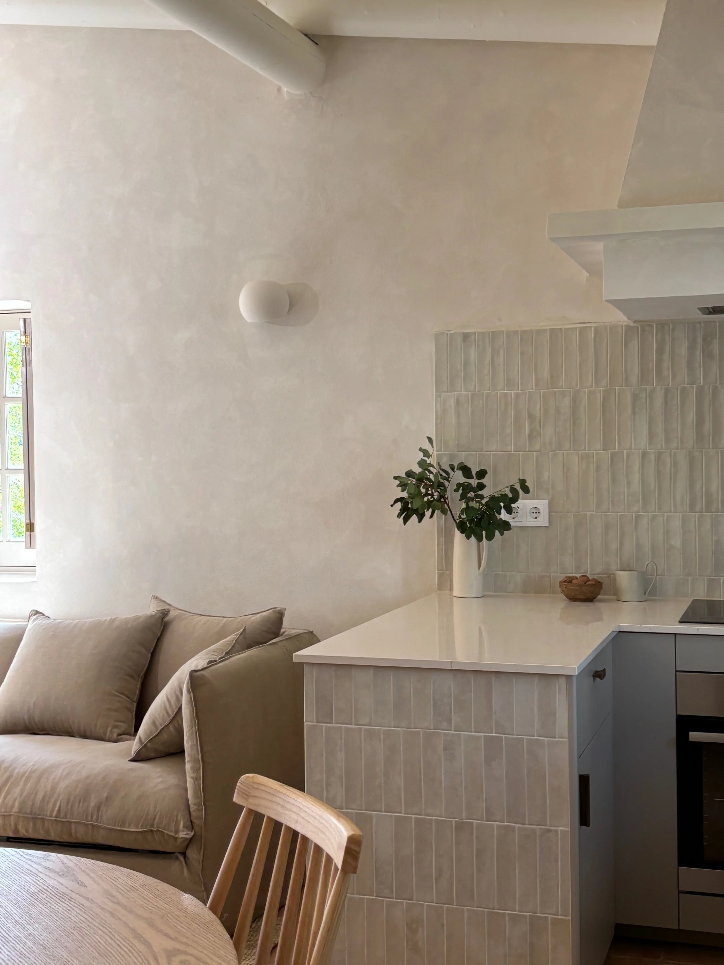 Interior of a modern living space showing a beige sofa with cushions, a partially visible wooden chair, and a white kitchen counter with a vase of green foliage, a bowl, and a mug. The wall behind features a textured finish and a window with window p