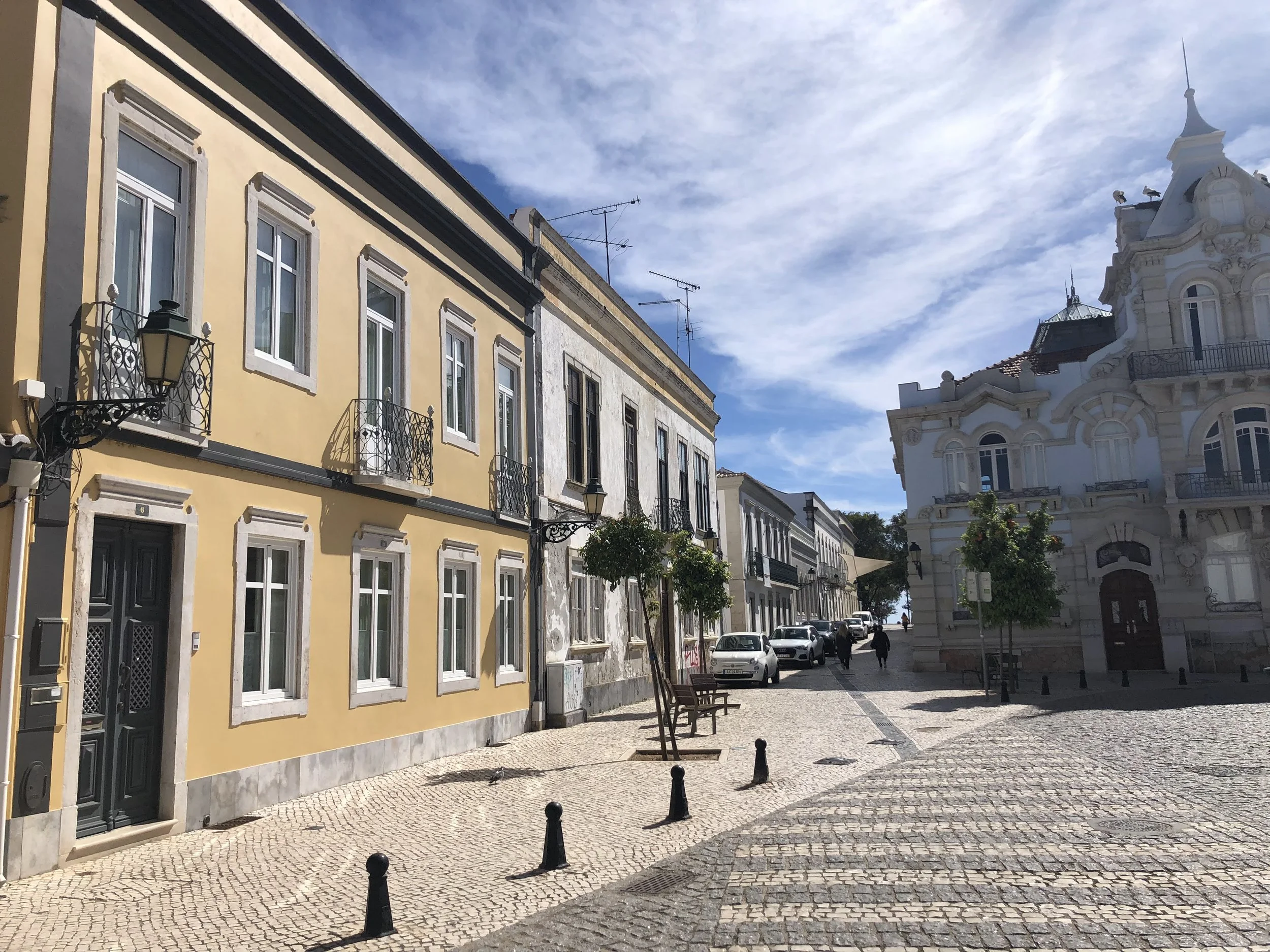 A Faro street scene with colorful historic buildings, cobblestone pavement, trees, parked cars, benches, and pedestrians under a partly cloudy sky.