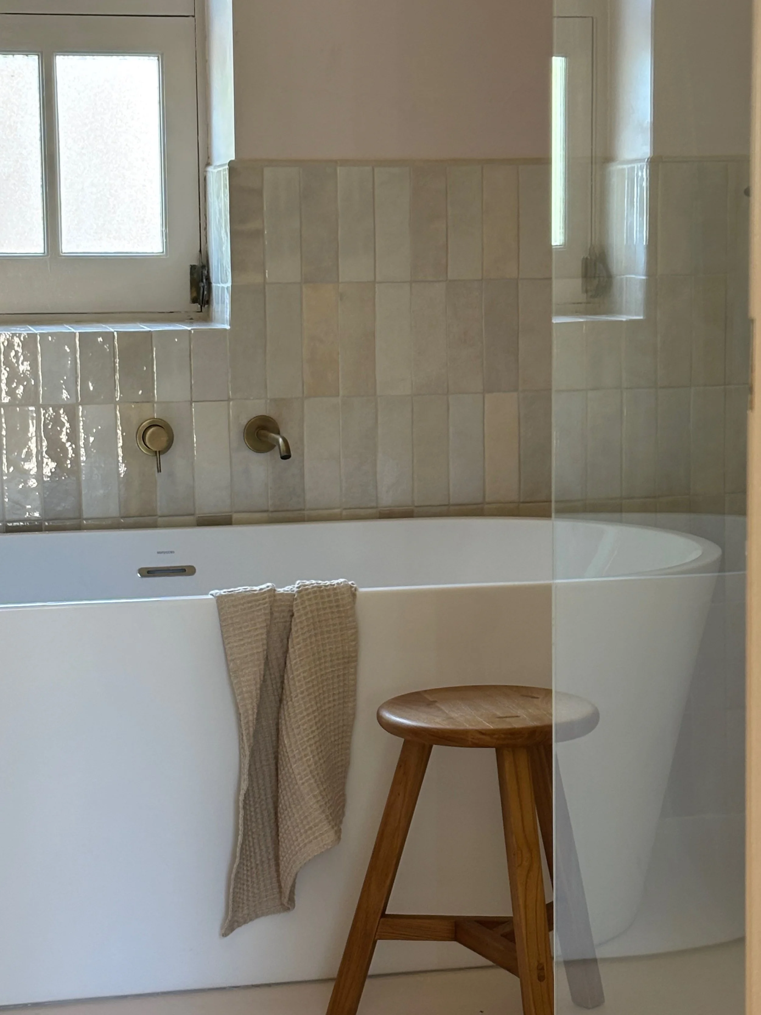 A modern bathroom with a white bathtub, beige tiled wall, a small wooden stool, and a window letting in natural light.