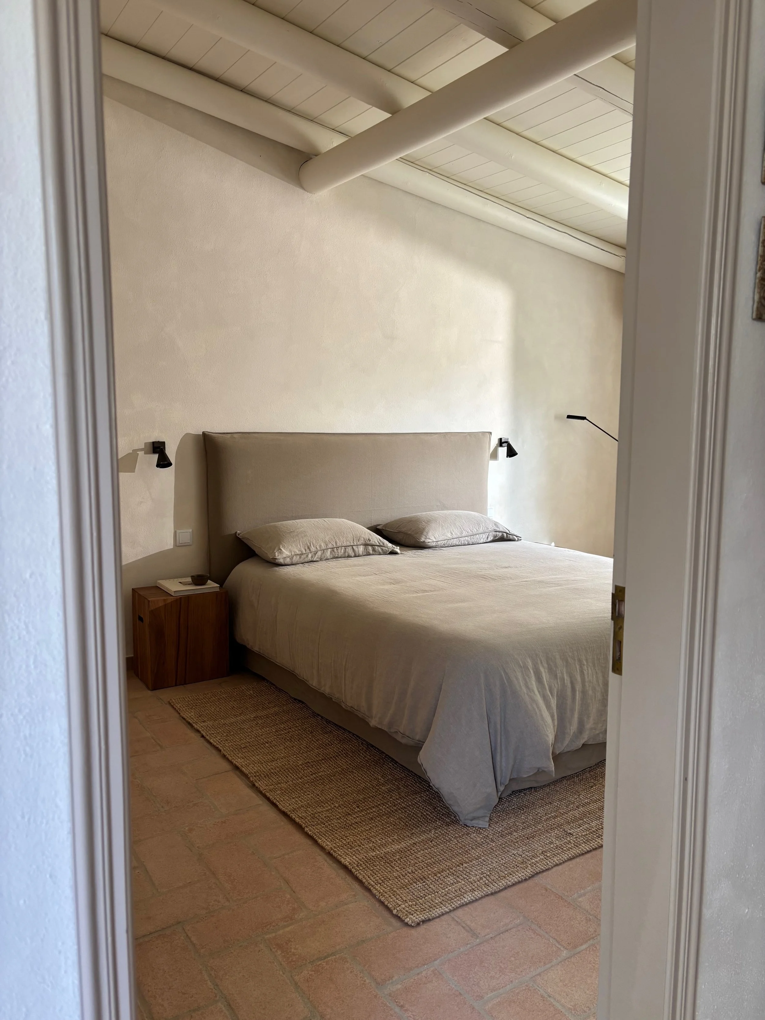 A minimalist bedroom with a neutral-colored bed and pillows, two black reading lamps on the beige wall, a wooden nightstand with a book and a small bowl, a textured rug, and a sloped ceiling with exposed beams.