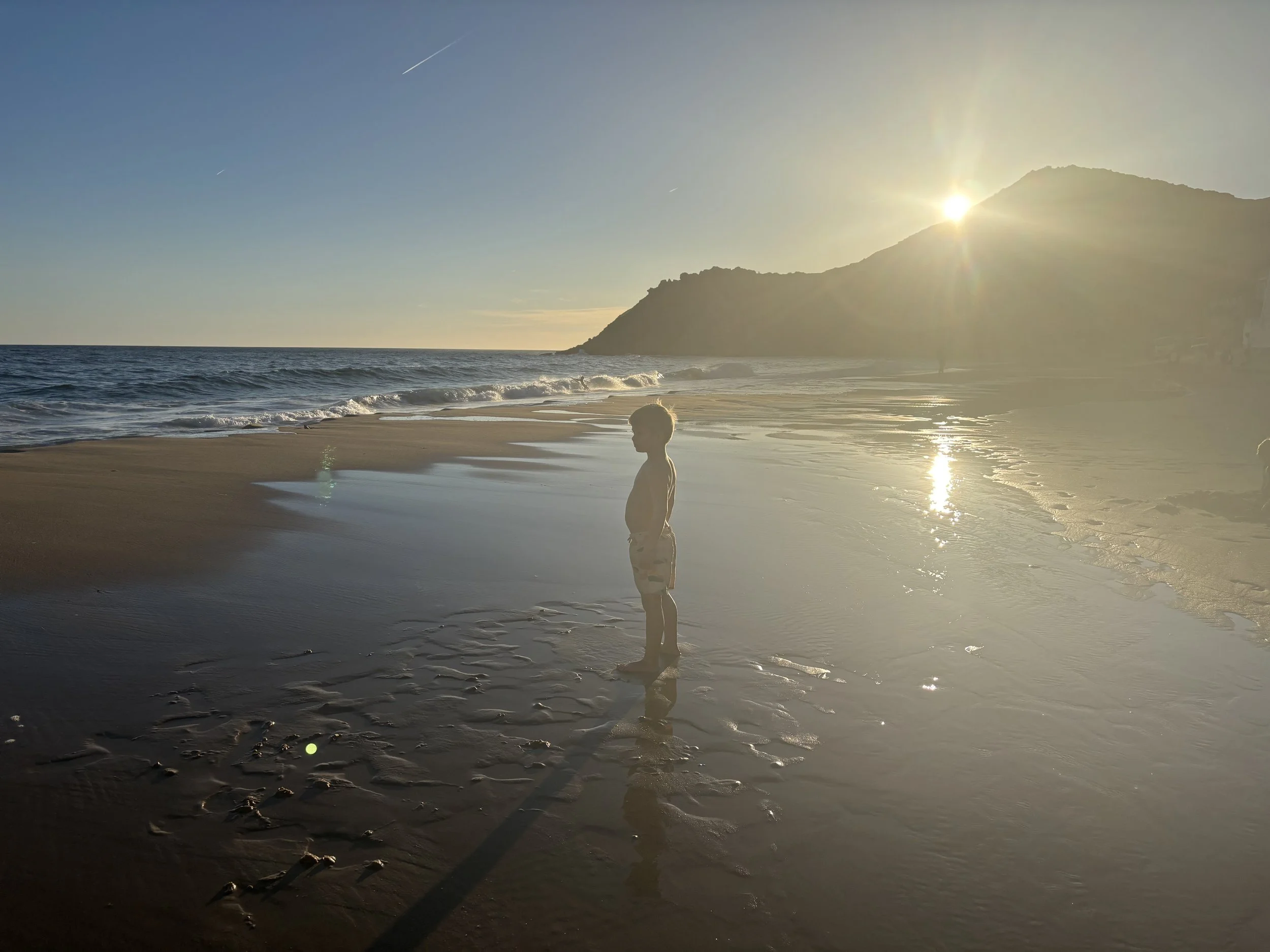 A young boy standing at the edge of the ocean during sunset, with the sun partially hidden behind a mountain, casting a warm glow on the beach and reflecting on the wet sand.