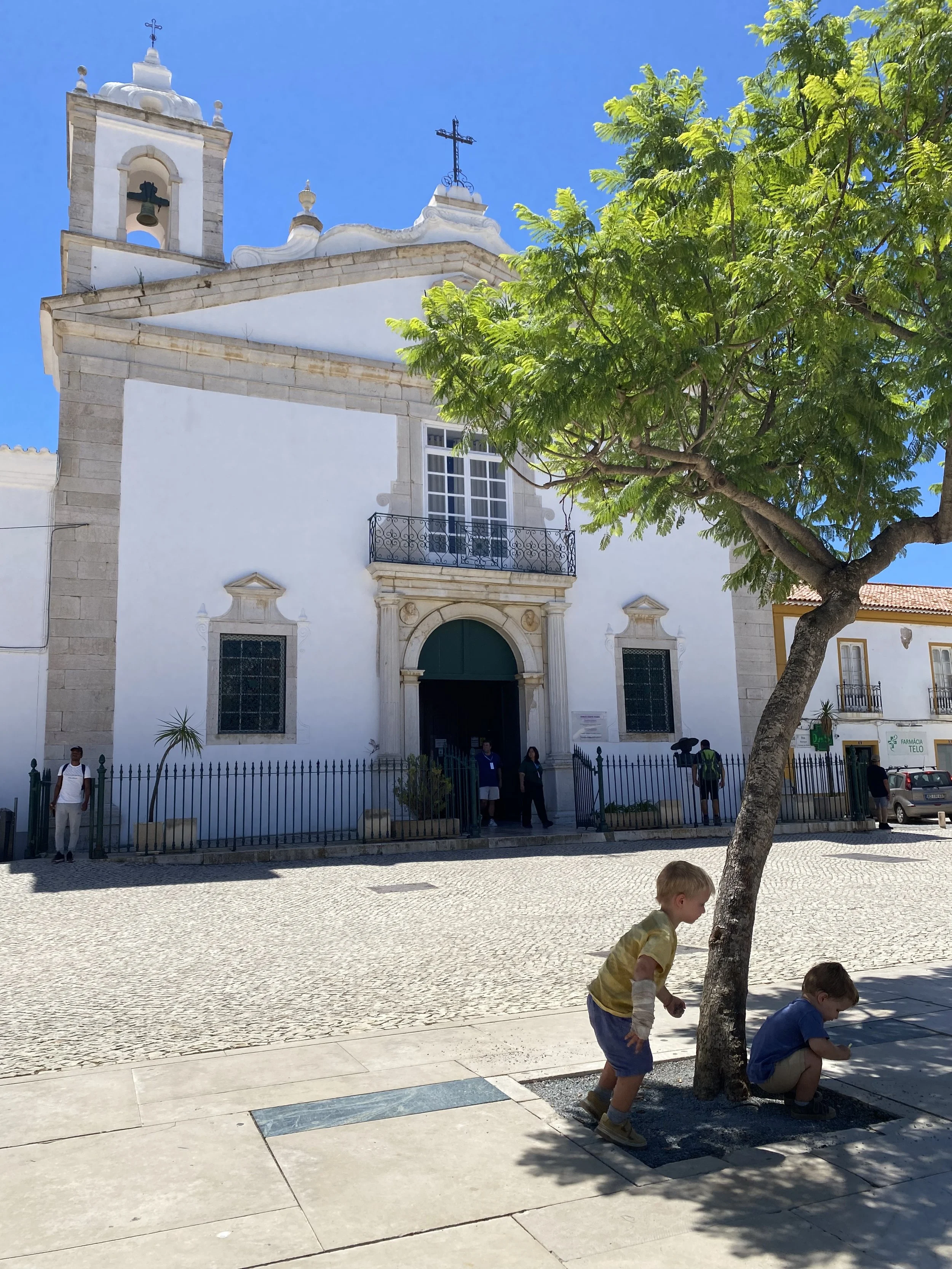 People standing in front of a white church with a bell tower, iron balcony, and crosses on top, under a bright blue sky, with children playing near a tree in the foreground.