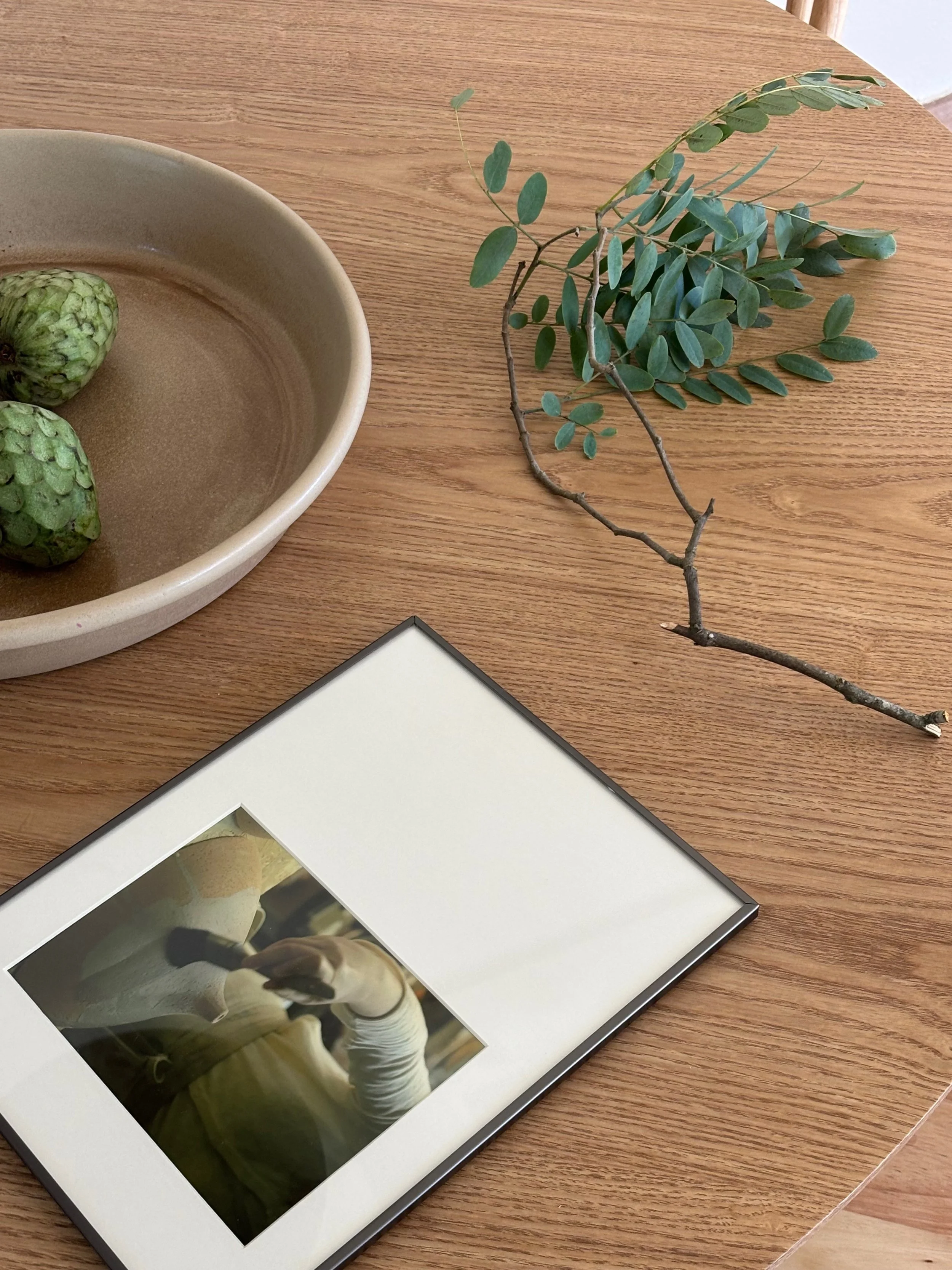 A wooden table with a beige bowl containing three green noni fruits, a sprig of green leaves, and a framed photo.