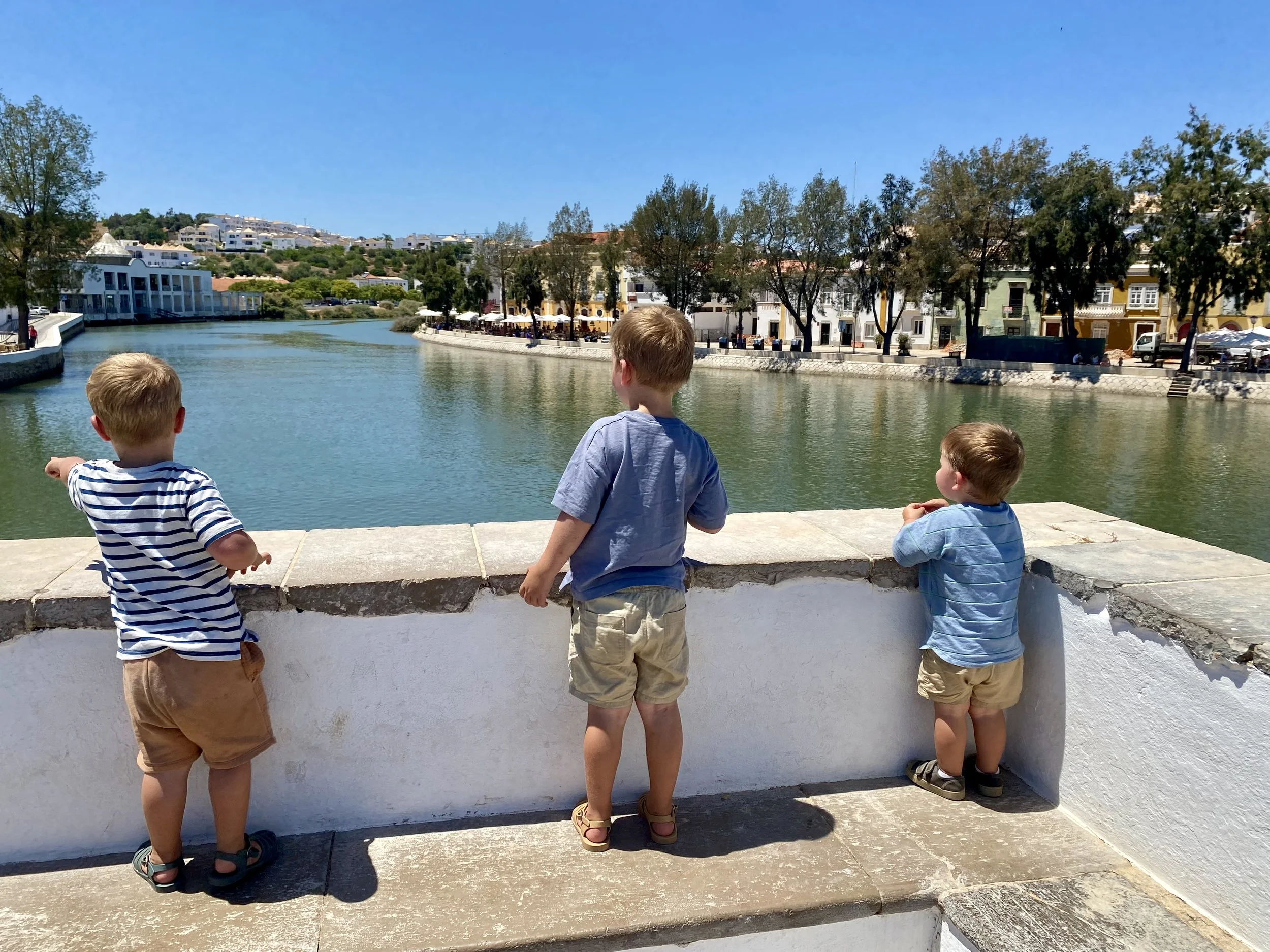 Three young boys standing on a stone ledge overlooking a river with houses and trees in the background on a sunny day.