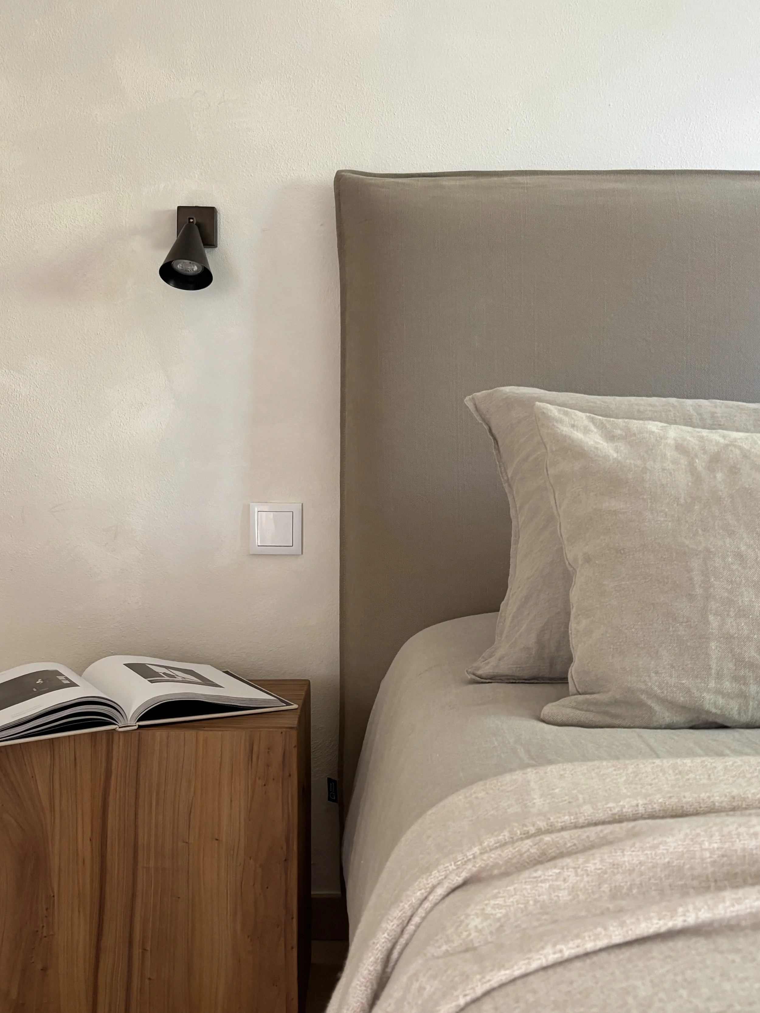 Close-up of a bedroom corner with a beige upholstered headboard, matching beige pillows, a wooden nightstand with an open book, a white wall, and a black wall-mounted reading light.