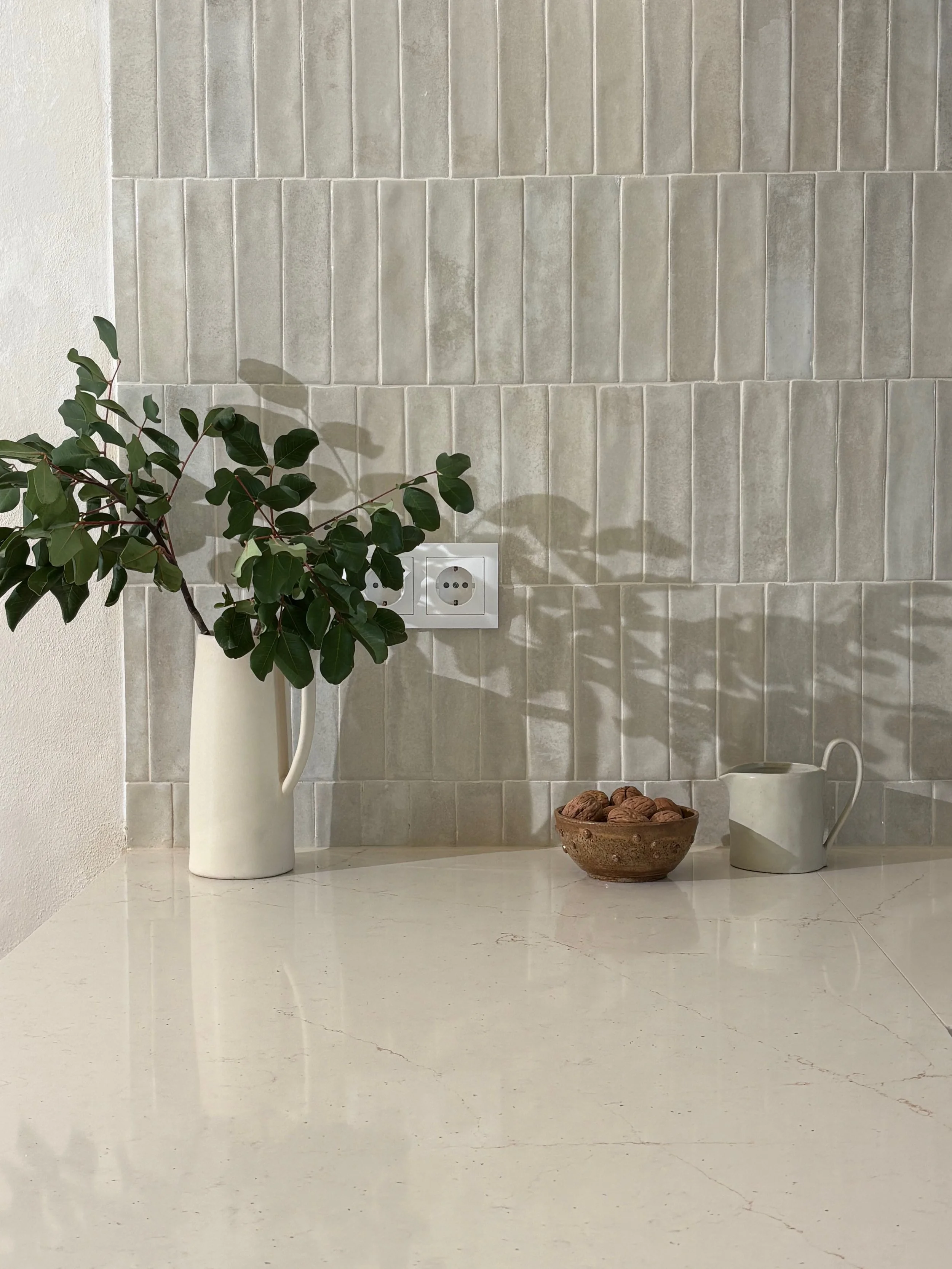 A white countertop with a green leafy plant in a white vase, a bowl of walnuts, and a small green and white pitcher in front of a beige tiled wall.