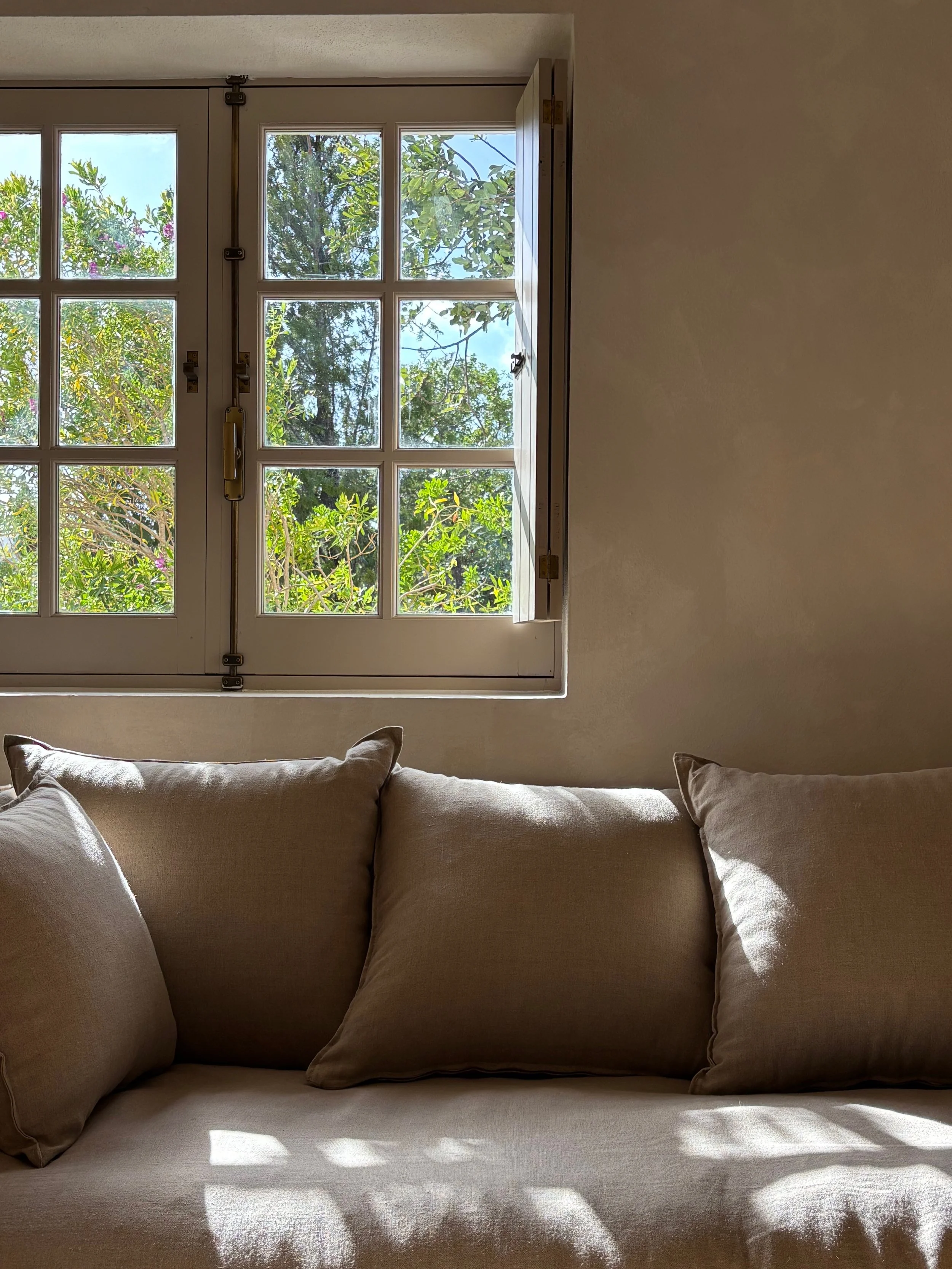 Sunlit beige sofa with pillows near a window overlooking green trees and blue sky.