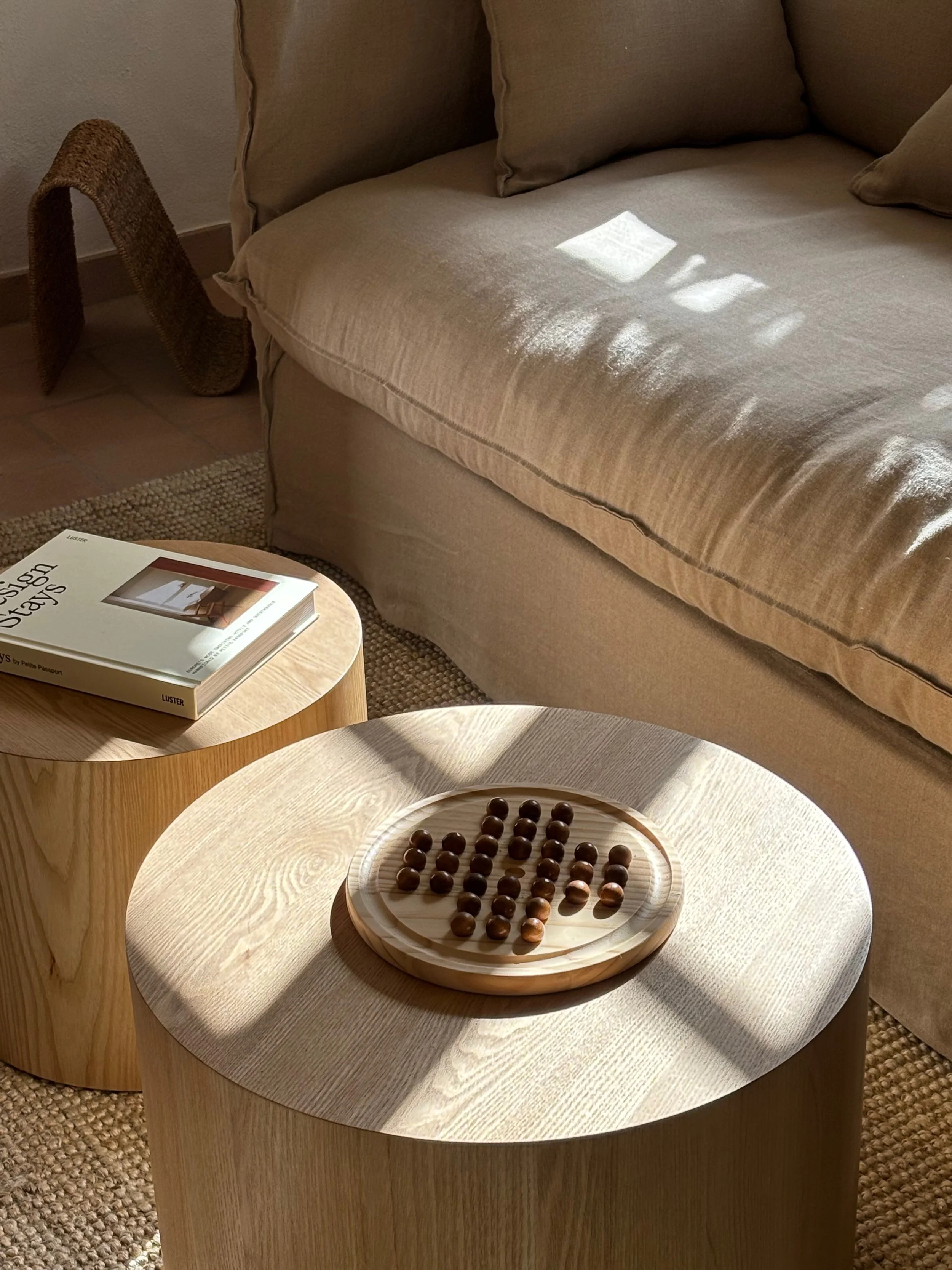 Living room corner with a beige sofa, two beige pillows, a small wooden side table with a book titled "Design Days" on it, and a round wooden coffee table with a wooden board game consisting of round wooden pieces. Sunlight creates shadows on the sof