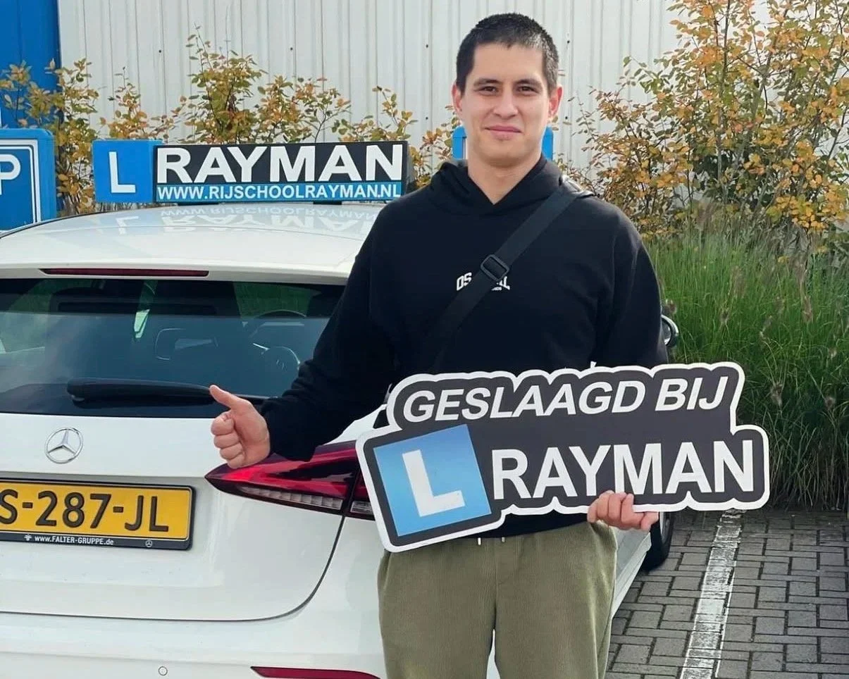 Man houdt een bord met de tekst 'Geslaagd bij Rayman' en een 'L' bord van een rijschool, achter een witte auto met een 'L' bord op het dak.