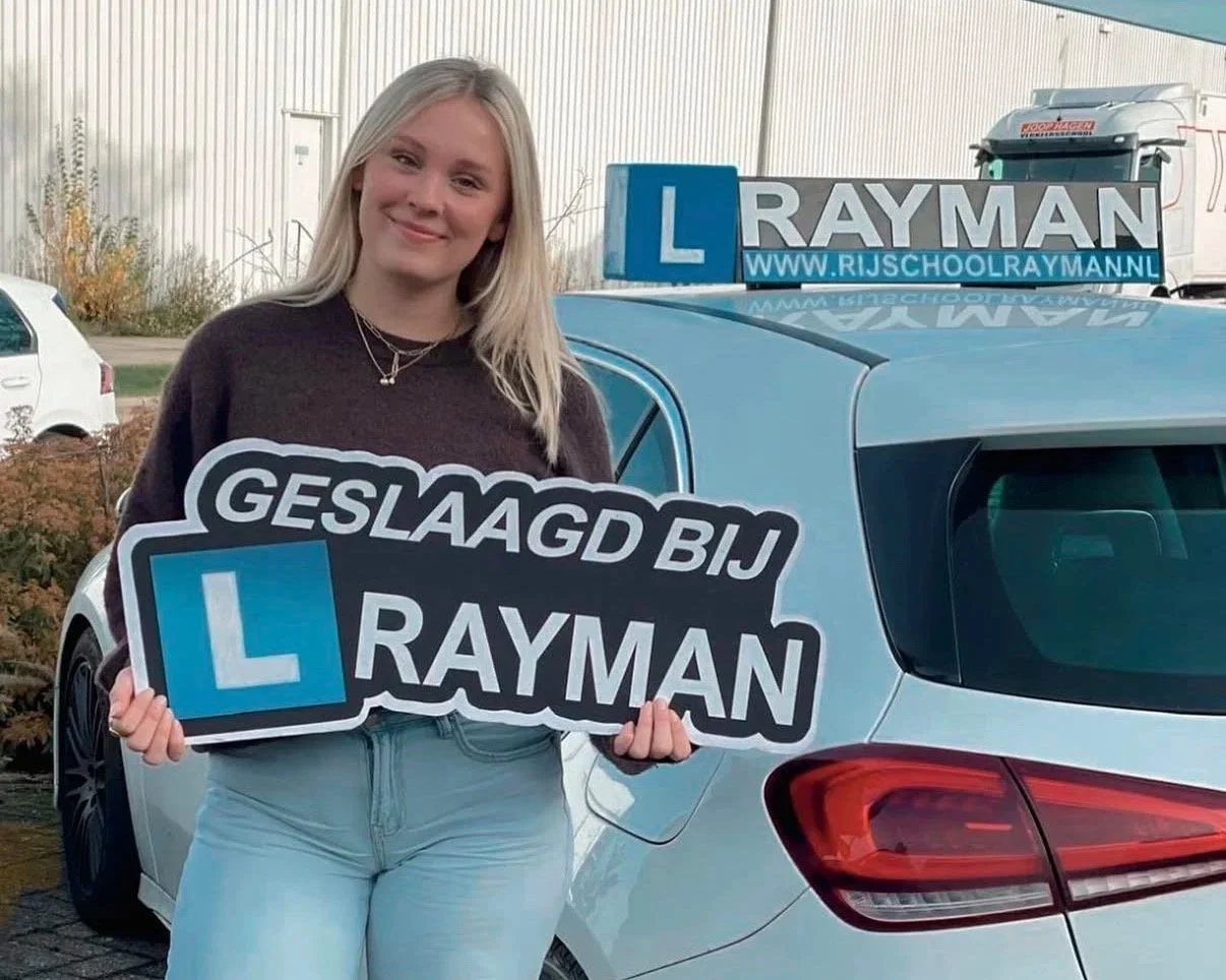 Vrouw houdt een bord met de tekst 'Geslaagd bij Rayman' en een auto met een 'L' rijschoolbord op het dak. 